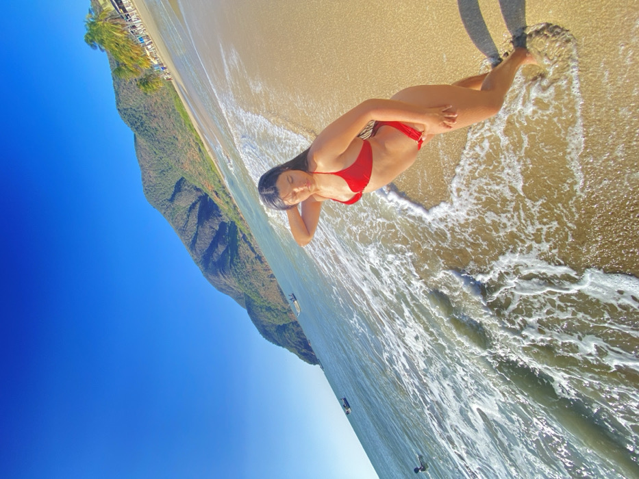 A woman in a red bikini lies on a sandy beach, partially submerged in the ocean's edge. Her head rests on her arm, eyes closed. A shadow of a person stands near her feet. Behind her is a mountain with lush vegetation. The sky is bright blue. The perspective is an extreme wide-angle shot, tilted and capturing the scene from a low vantage point. Waves wash over the sand.