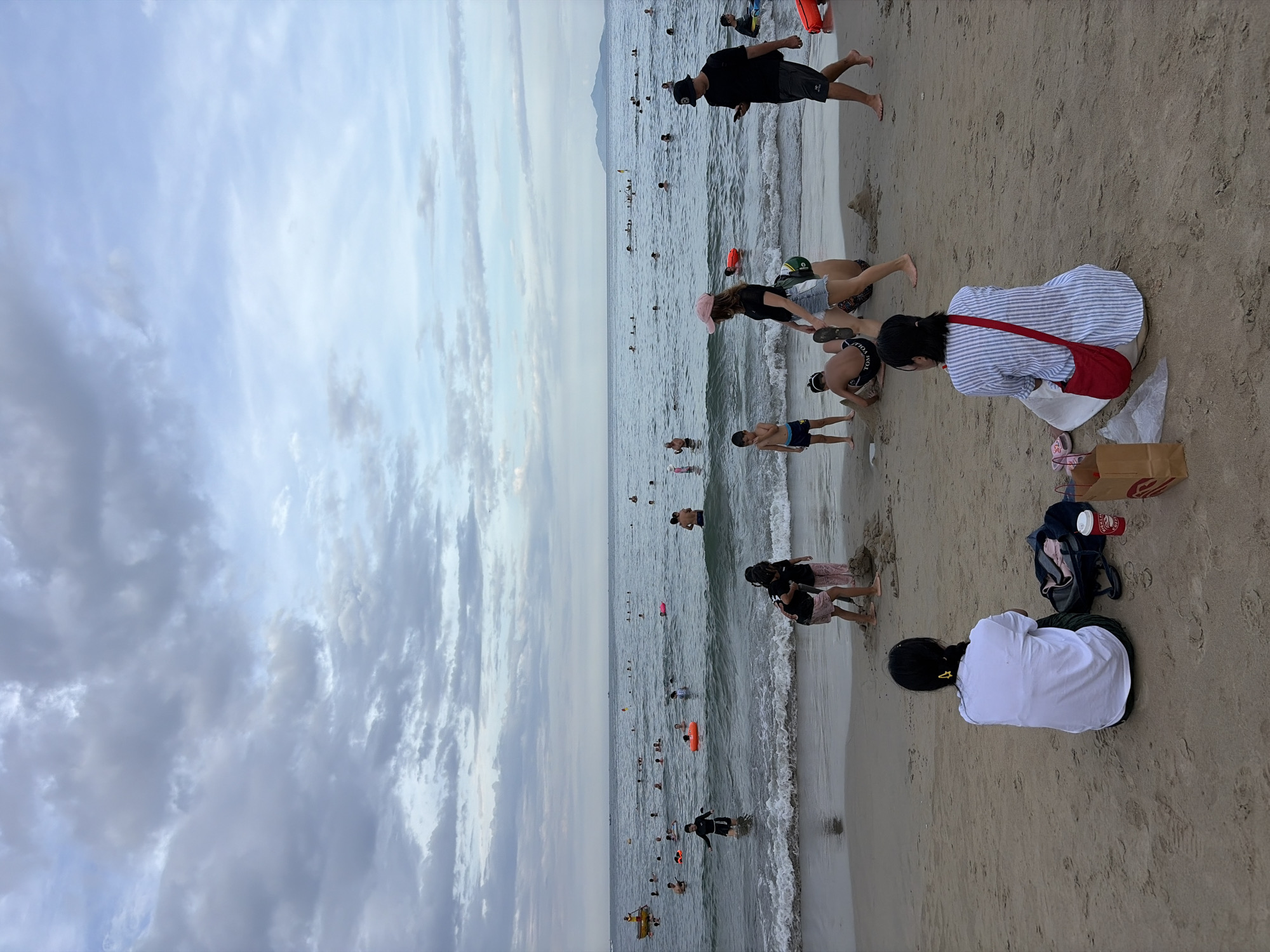 The image depicts a crowded beach in Da Nang, Vietnam, with the ocean in the background and people engaged in various activities. The sky is overcast with a mix of grey and blue clouds. The sea is moderately wavy, with numerous individuals swimming and playing in the water. Several people are visible on the sandy beach, including children building sandcastles and adults relaxing.
In the foreground, two women are seated on the sand, facing away from the camera. The woman on the left wears a white t-shirt and dark pants, with a black bag and a red cup beside her. The woman on the right is dressed in a blue and white striped shirt and carries a red bag over her shoulder.
Further back, several groups of people are interacting. A man in a black shirt and shorts is walking on the beach, looking at his phone. Two children are playing near the water's edge, and a man in a dark hat is also present. In the water, a group of people are swimming, with some using inflatable rings.
The beach environment suggests a warm day, likely in the late afternoon or early evening, given the cloud cover and the overall lighting. The presence of numerous people indicates a popular tourist destination or a local gathering spot. The shallow waves suggest it is a safe area for swimming.