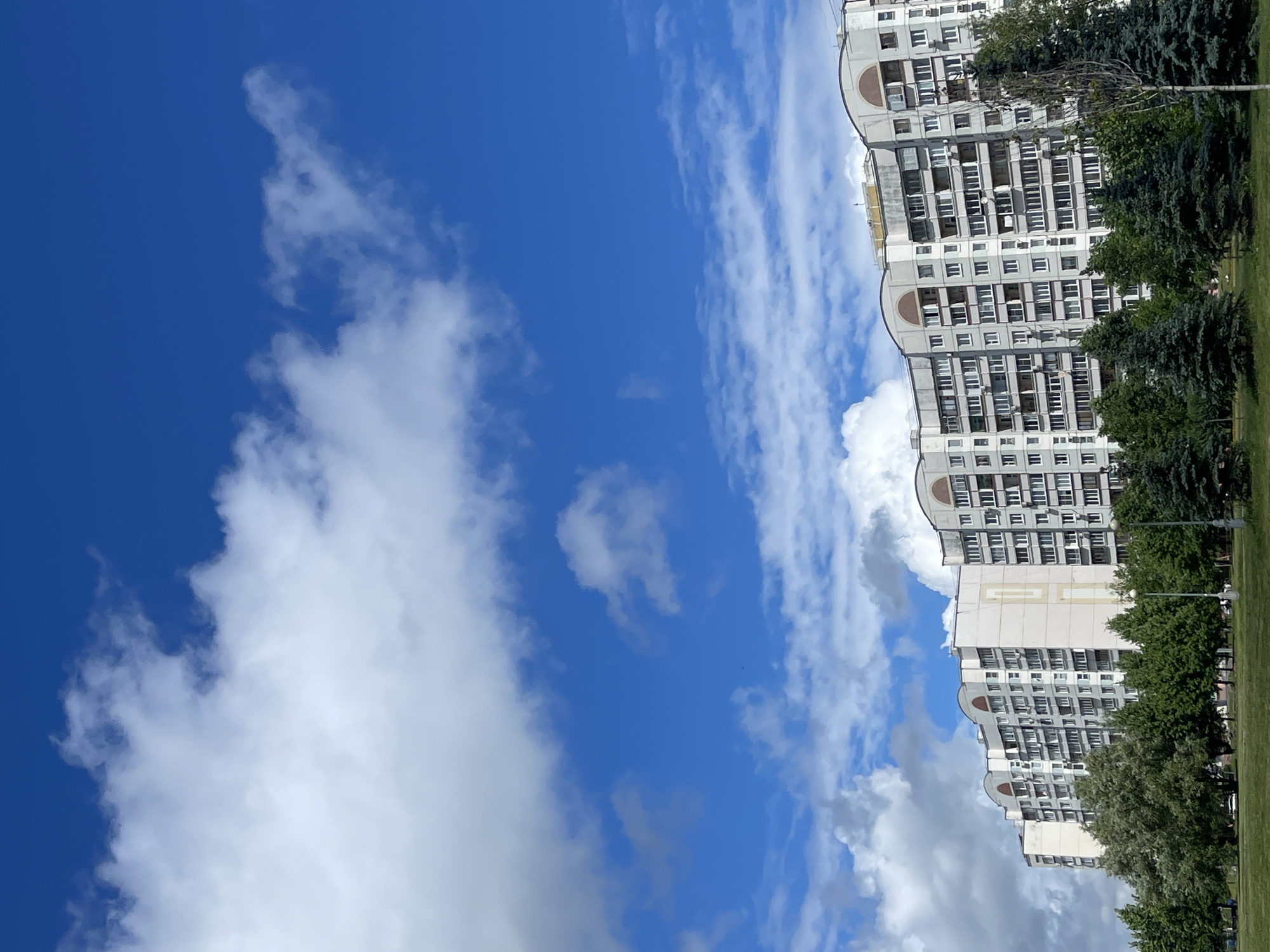 The image shows a residential apartment building in a suburban area of Moscow, Russia. The building is a multi-story structure with a light-colored facade and arched architectural elements at the top. There are numerous windows and balconies on the building, indicating it is an apartment complex. In front of the building, there is a line of various trees, including deciduous and coniferous varieties, and a well-maintained green lawn. The sky is bright blue with scattered white, fluffy clouds. The overall impression is a typical urban residential scene on a clear day.