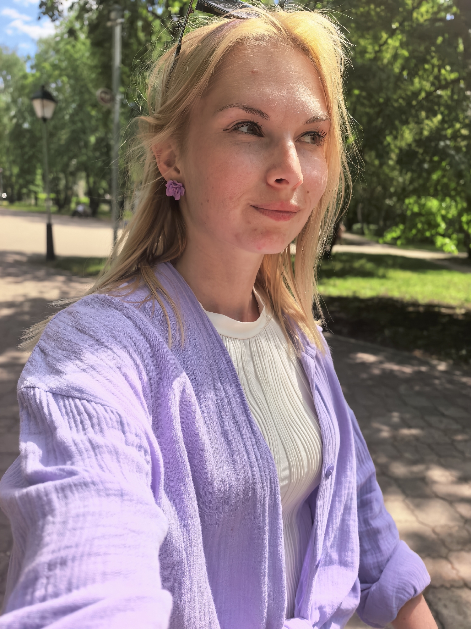 A young woman stands outdoors, seemingly in a park or green space, likely in Sterlitamak, Russia, based on the prompt. She is fair-skinned with blonde hair, wearing a white top and a light purple open-front shirt or cardigan. She has purple floral earrings. Her expression is thoughtful, and she is looking slightly off-camera to her right. The background features trees, possibly a street lamp, and dappled sunlight on a paved surface.