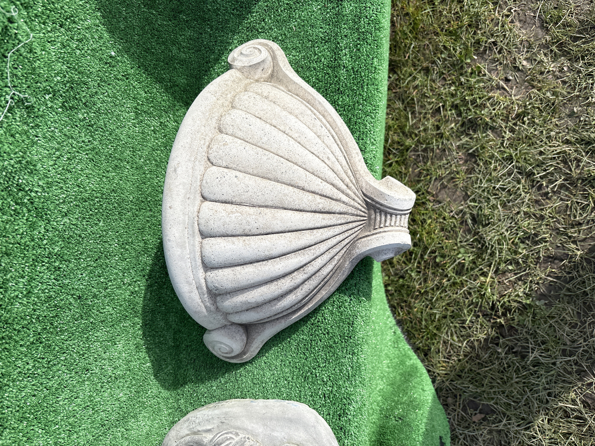 The image shows a grey, shell-shaped architectural ornament, possibly made of concrete or stone, resting on a square of artificial green turf. To the right of the turf, there is a patch of bare earth with sparse vegetation. The photograph was likely taken outdoors in daylight. The location in the city of Chirk, United Kingdom is not discernible from the image. There are no people visible in the image.