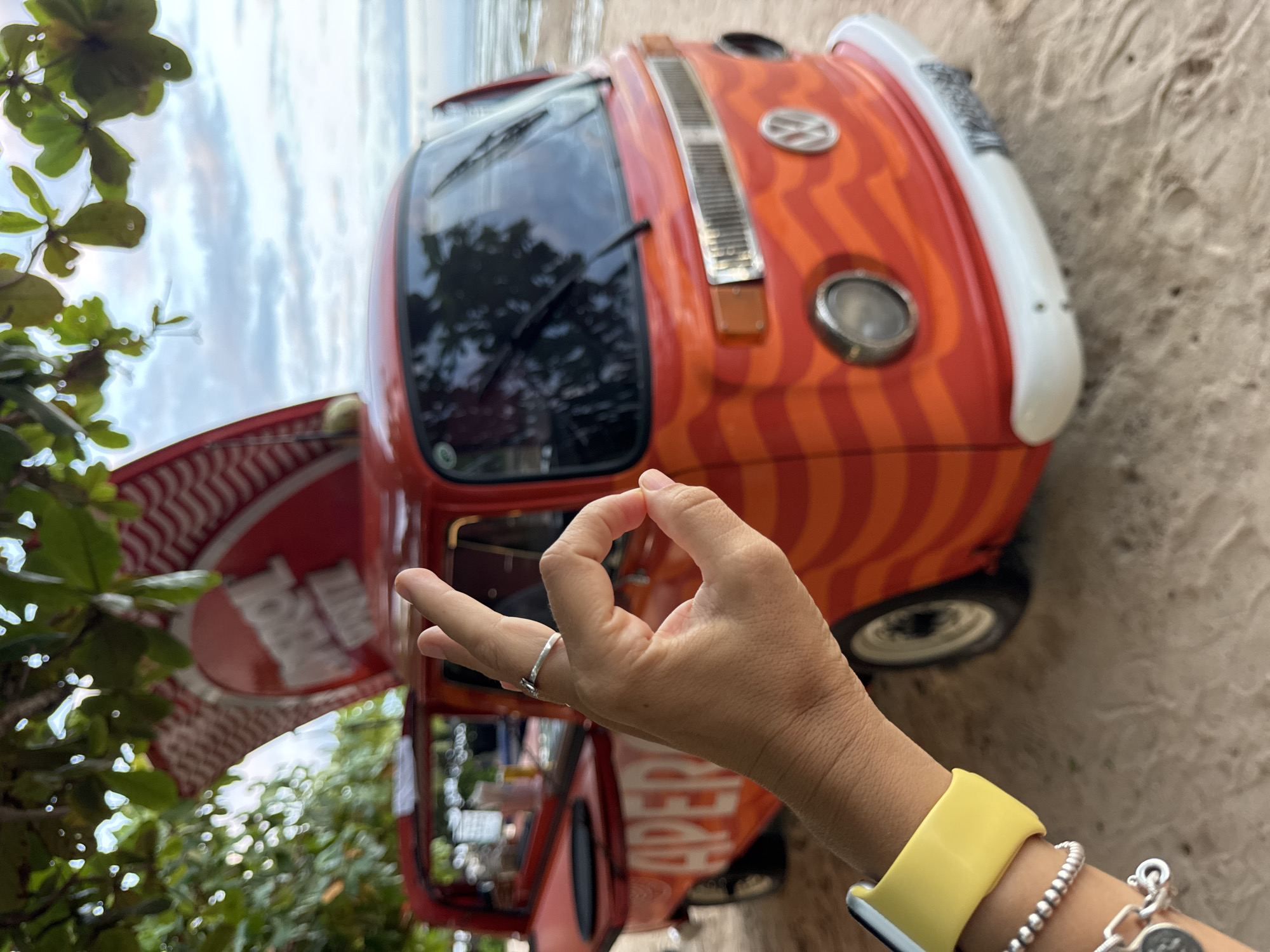 A photograph captured at an unspecified location in Kuta Selatan, Indonesia, features a partially visible red and white Volkswagen van parked on a sandy surface, likely a beach. The van has an open side hatch, revealing an "Aperol Spritz" logo. Dominating the foreground is a female hand, adorned with a silver ring on the middle finger, making an "OK" gesture. The wrist wears a yellow sports watch band and several silver bracelets. Vegetation and a cloudy sky can be seen in the background.