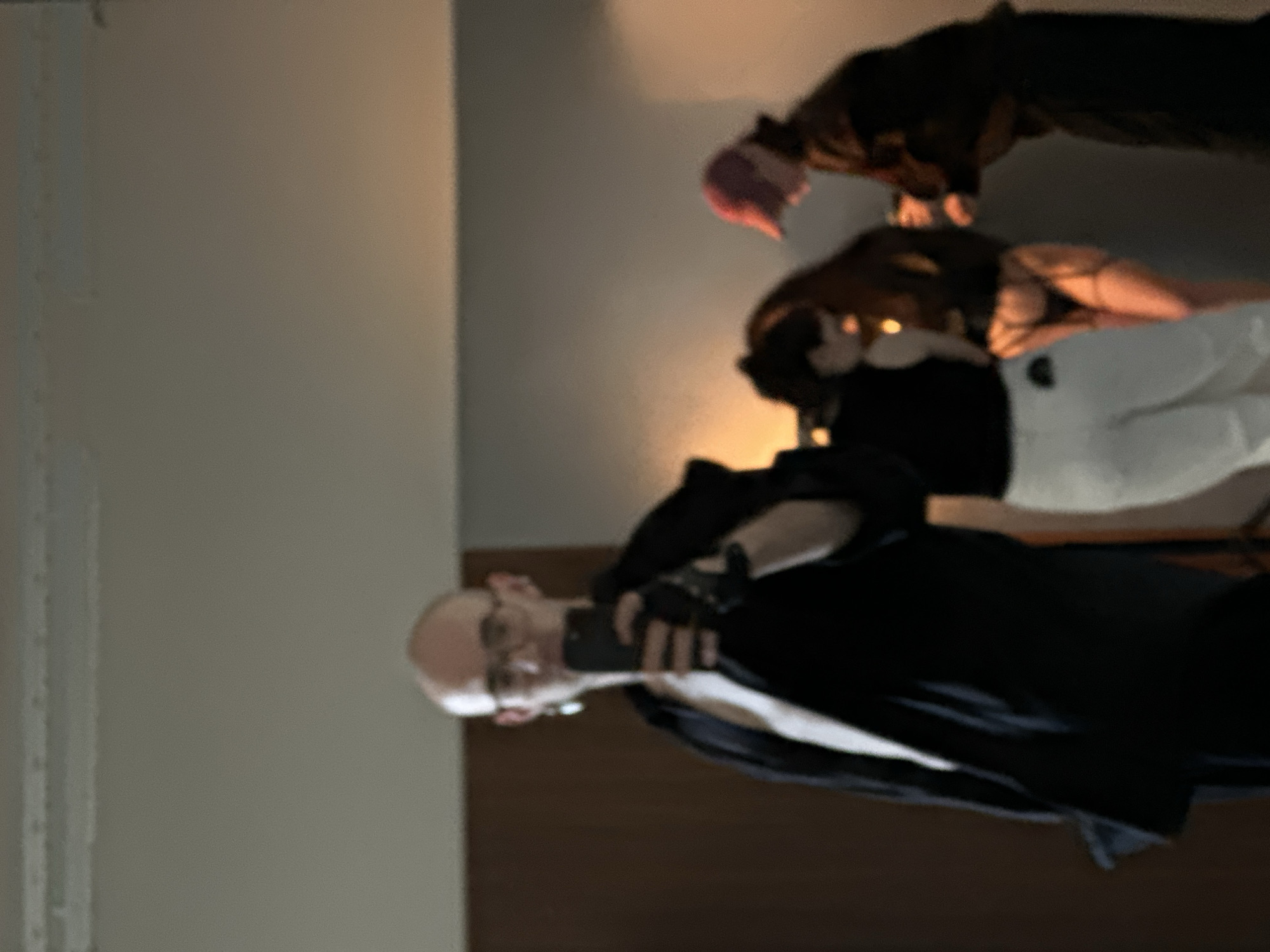 The image depicts three individuals indoors. One person, bald and wearing glasses, holds a phone and is dressed in black with a white garment underneath. Another person wears a black top and white pants. A third individual with pink hair is also present. The location is potentially Kazan, Russia, but this is not confirmed by image content alone. There is a light brown wall.