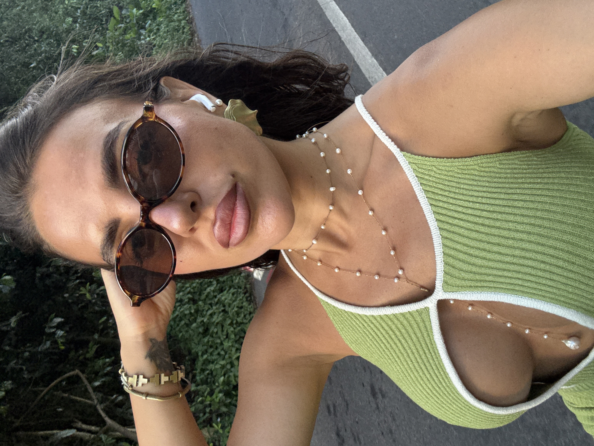 A woman is taking a selfie outdoors. She is wearing a green knitted top with white trim, brown sunglasses, and jewelry including a pearl necklace and gold bracelets. She also has a white earbud in her ear. The background shows greenery and a paved road, which may suggest that the location is in Kuta Selatan, Indonesia.