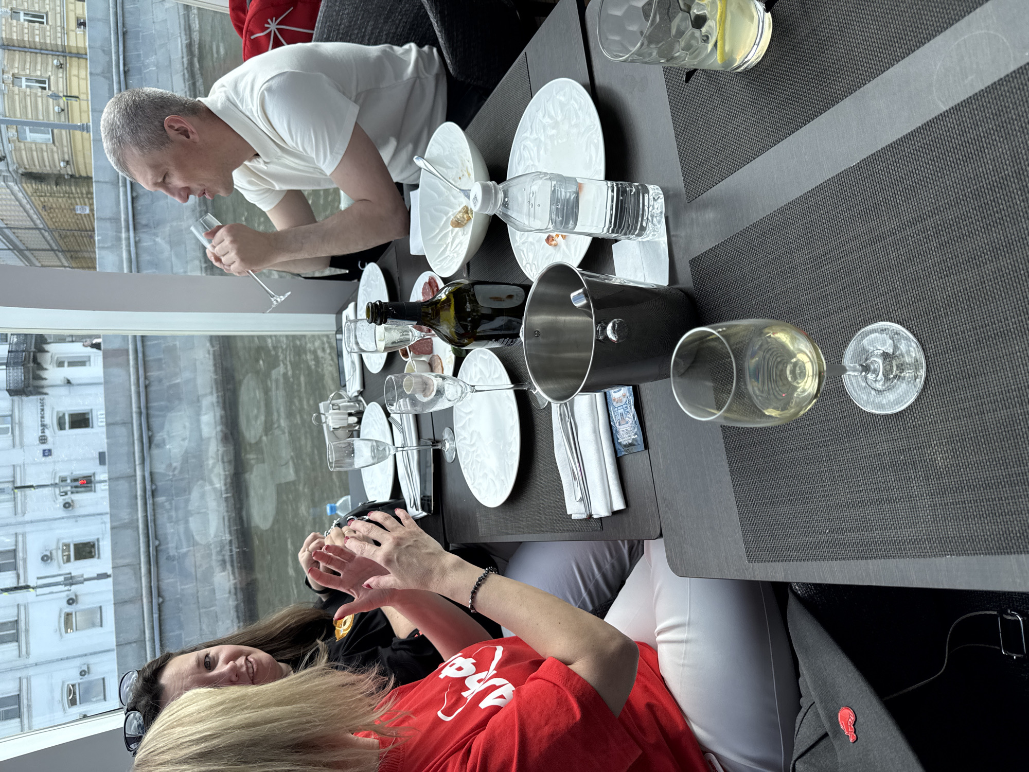 The image shows a group of people dining at a restaurant. A man in a white shirt is holding a wine glass, seemingly examining its contents. A woman in a red shirt is sitting across from him, gesturing with her hands while speaking to someone out of frame. The table is set with wine glasses, plates, cutlery, a wine bottle, and a bottle of water. There are also remnants of a meal on some of the plates, suggesting that they have already eaten. Through the window, a view of a building with the word "АРБАТСКАЯ" on it can be seen, indicating the location is likely in the Arbat district of Moscow, Russia. A river also runs past the building.