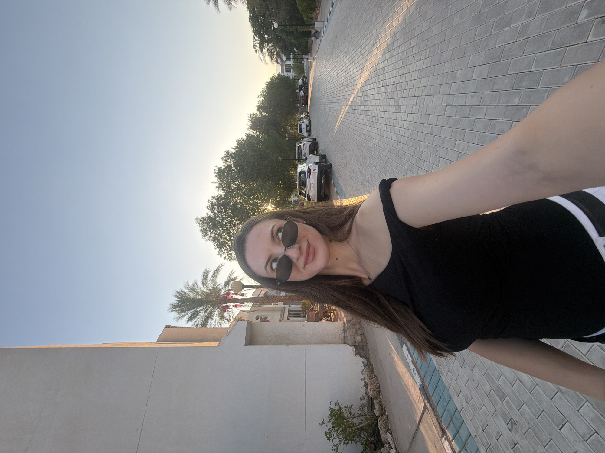 A young woman with long brown hair, wearing sunglasses and a black off-the-shoulder top, poses for a selfie on a paved sidewalk in Sharm El Sheikh, Egypt. The sun, low in the sky, casts long shadows and illuminates the scene with warm golden light. To the left, a white building with a textured facade stands adjacent to the sidewalk, with a small green bush and some rocks at its base. Across the street, a row of parked cars is visible, with lush green trees lining the background. The sky is a clear, pale blue. The woman is looking away from the camera, towards the right side of the image, with a slight smile.