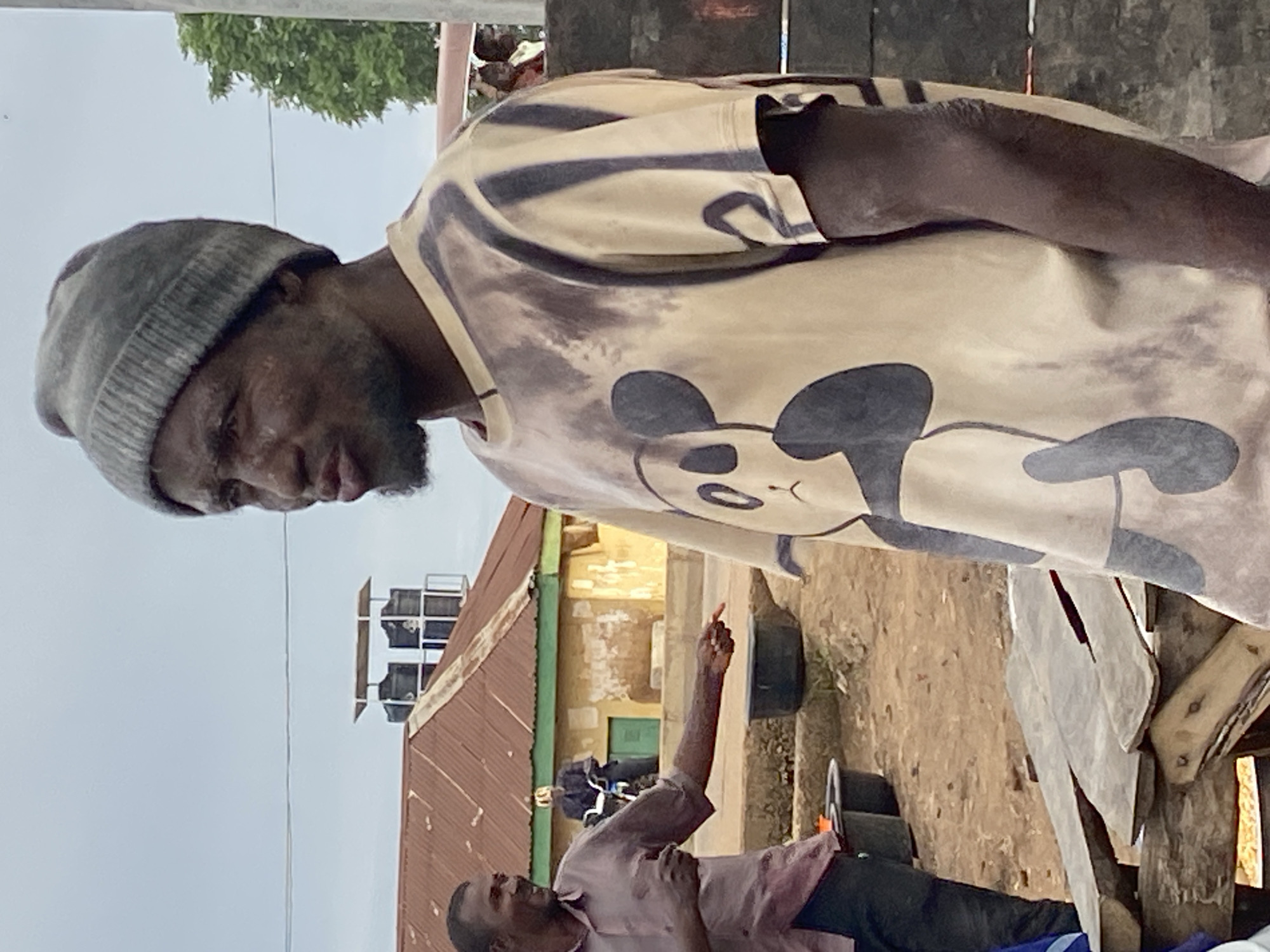 In Bukuru, Nigeria, a man wearing a grey knitted beanie and a tan t-shirt with a panda design is visible in the foreground. He has dirt on his face and appears to be speaking. Behind him, another man in a maroon shirt is gesturing with his hand, and a person on a motorcycle is visible further back. In the background, there are buildings with corrugated metal roofs, and the ground is dirt. The scene appears to be outdoors in a somewhat dusty environment.