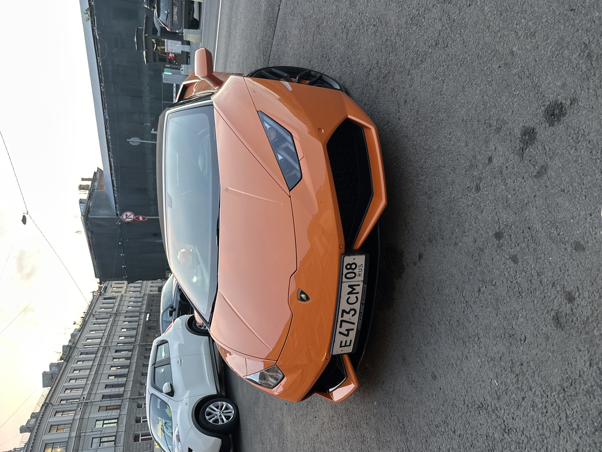 The image shows the front of an orange Lamborghini Huracan parked on a street in Saint Petersburg, Russia. The car's license plate is visible, reading "E473 CM 08 RUS". To the left of the Lamborghini is a white Nissan Note and a section of a large, gray building with numerous windows, indicating an urban environment. The street surface is asphalt with visible patches and stains. The sky is overcast or the photo was taken during twilight, casting a soft light on the scene. A dark building with scaffolding is behind the Lamborghini. A street light is visible in the upper left portion of the frame.