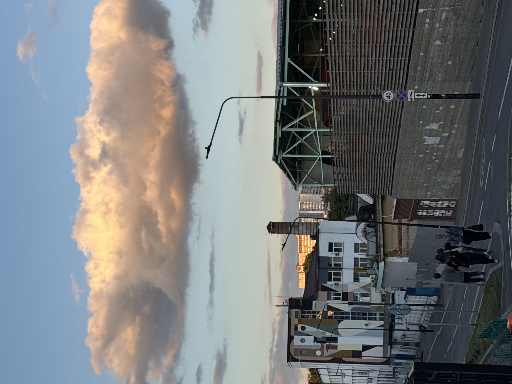 The image shows a street scene in Moscow, Russia. The photo is oriented vertically. On the left side of the frame is a large, light-orange cloud in a blue sky. A tall, industrial-looking building is on the right, with a corrugated metal facade and a green steel framework. In front of this building is a streetlight and a road with a "40" speed limit sign and other street signs. Further down the street are residential buildings with windows and geometric patterns on their facades. A group of people is walking on the sidewalk.
