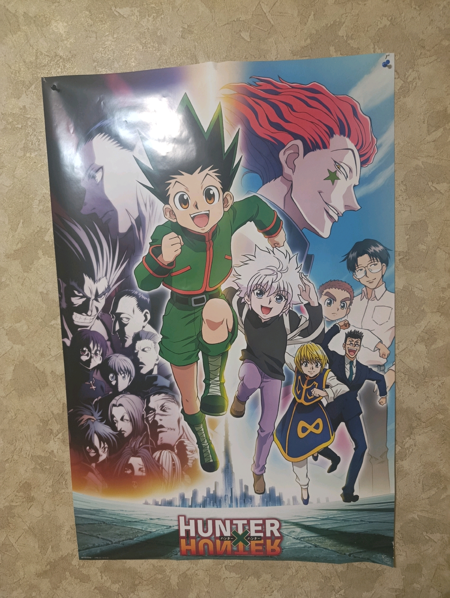 The image shows a poster of the anime series "Hunter x Hunter" hanging on a textured, beige wall. The poster features several characters from the show, including Gon Freecss, Killua Zoldyck, Hisoka Morow, and others. The characters are depicted in a dynamic, action-oriented pose. The poster is attached to the wall with at least two pushpins, one visible in the top left corner and another in the top right corner. The lighting suggests an indoor environment with a light source casting some shadows. The location in the city of Horishni Plavni, Ukraine, is not discernible from the image itself.