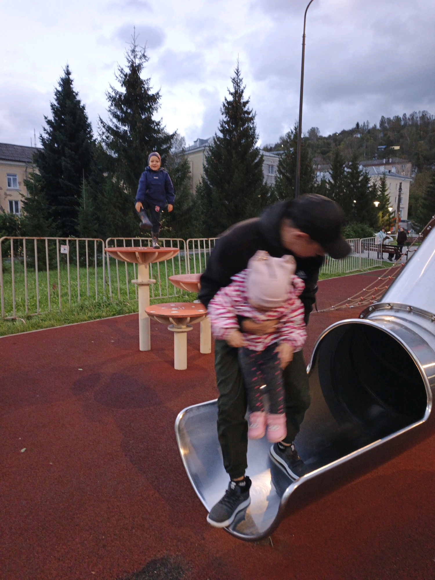 In Zlatoust, Russia, on a playground with a red rubberized surface, a person in a black hoodie and baseball cap holds a young child wearing a pink and white jacket and a light pink hat. The child is positioned in front of the adult, facing the viewer, with their arms around the adult's neck. Both are on a large, silver metal slide that curves to the right. To the left, a child in a blue jacket and grey pants stands on a mushroom-shaped playground structure, looking towards the viewer. In the background, a white metal fence encloses the playground, beyond which are trees and buildings, including a light-colored residential building. The sky is overcast and grey, with some clouds visible, and a street lamp stands in the upper right quadrant of the image.