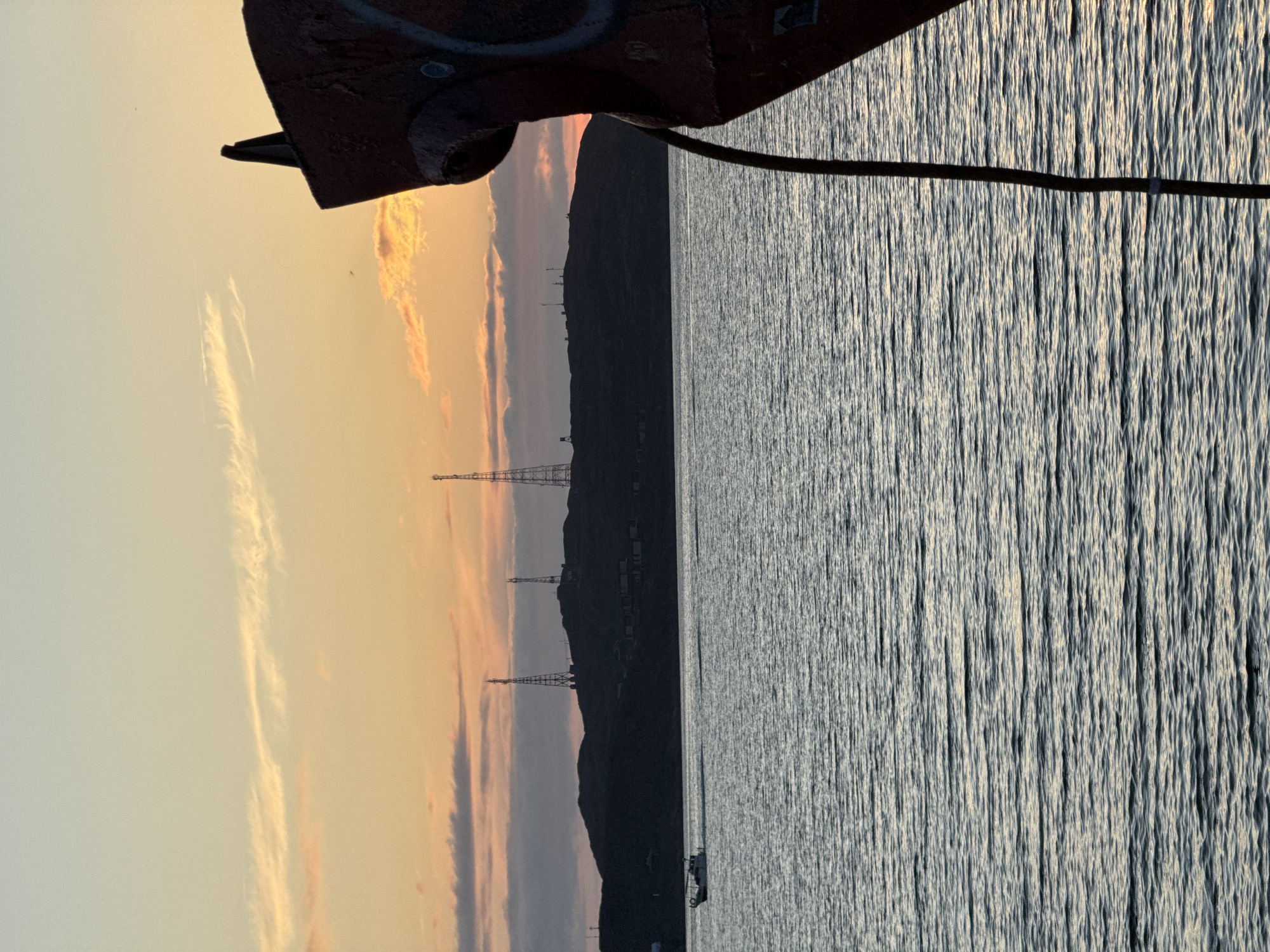 This image depicts a view of the Teriberka Bay in Teriberka, Russia, during sunset. The sky is a gradient of pale blue and orange, with wispy clouds. In the distance, a silhouetted hill is visible, topped with communication towers and some buildings. A small white boat is moving across the water. In the foreground, the rusted bow of a ship juts into the frame from the right, with a thick rope hanging down. The water is calm with ripples.