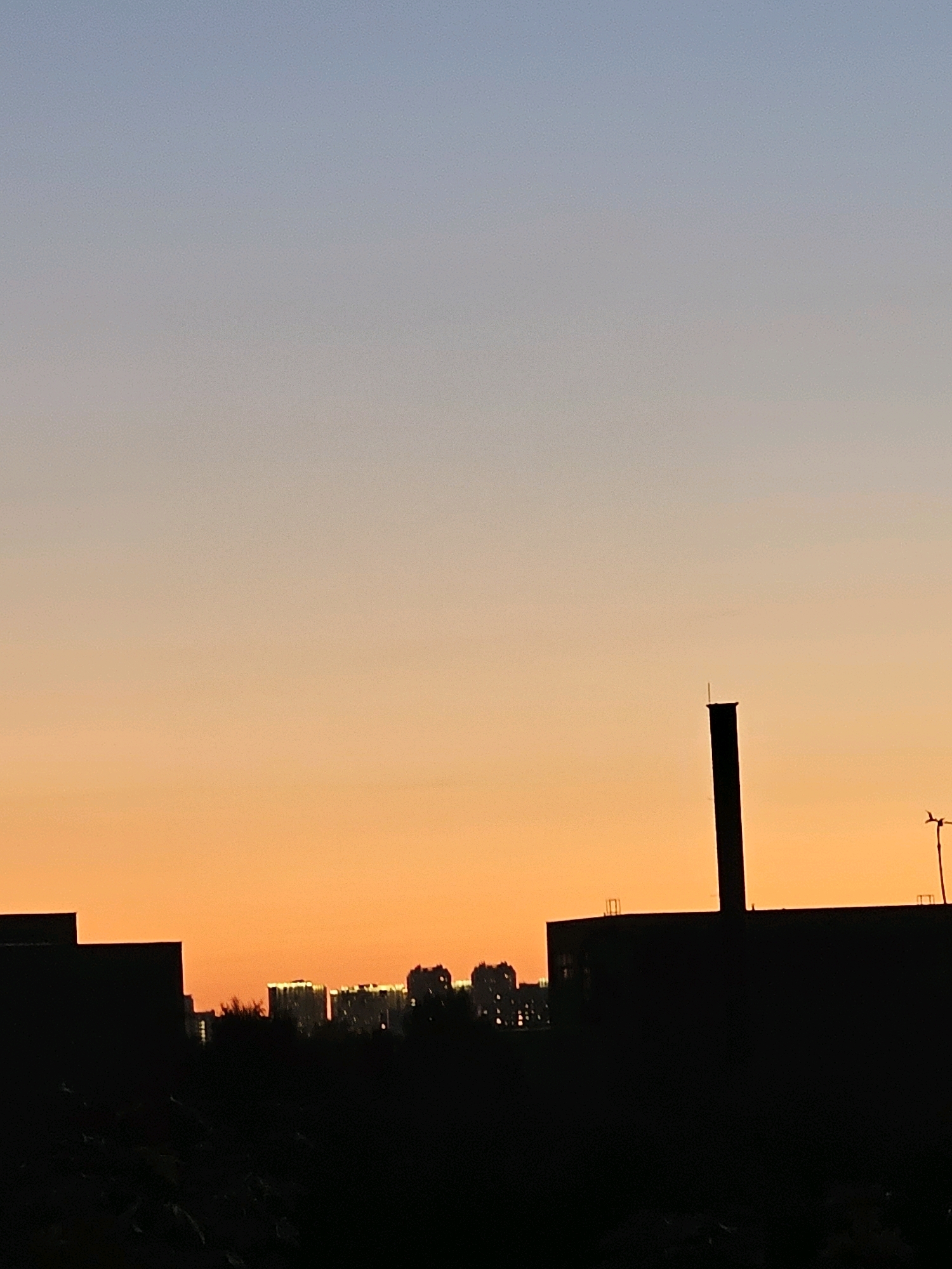 A silhouette of a city skyline is visible against a gradient sunset sky, transitioning from pale blue at the top to a vibrant orange near the horizon. The cityscape comprises several dark, angular structures, including what appears to be a tall, slender smokestack or industrial pipe in the center-right of the frame. In the distance, illuminated buildings suggest an urban area. The foreground is dominated by a dark, amorphous shape, likely trees or debris, obscuring the ground. The overall impression is of a twilight urban scene in Khimki, Russia. There are no people or discernible animals in the image.