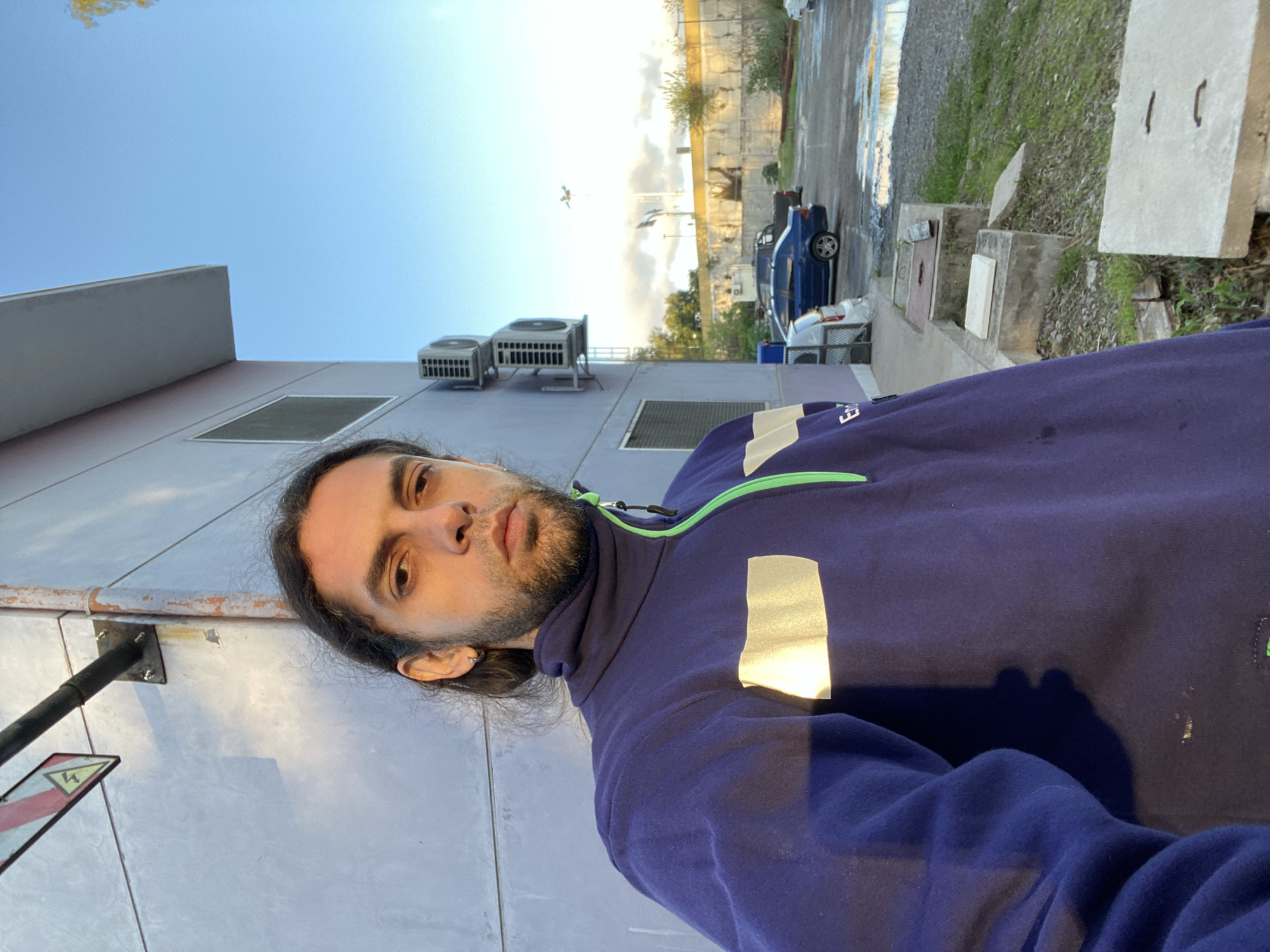 A medium-shot selfie depicts a man with long, dark hair tied back and a beard, wearing a navy blue sweatshirt with a light green zipper and reflective rectangles on the front. The background shows an exterior scene with a light blue building, air conditioning units, and a utility sign mounted on the wall. Beyond the building, a scene with parked cars, trees, and the sky is visible. The photograph does not provide sufficient information to confirm the specific location in Buenos Aires, Argentina.