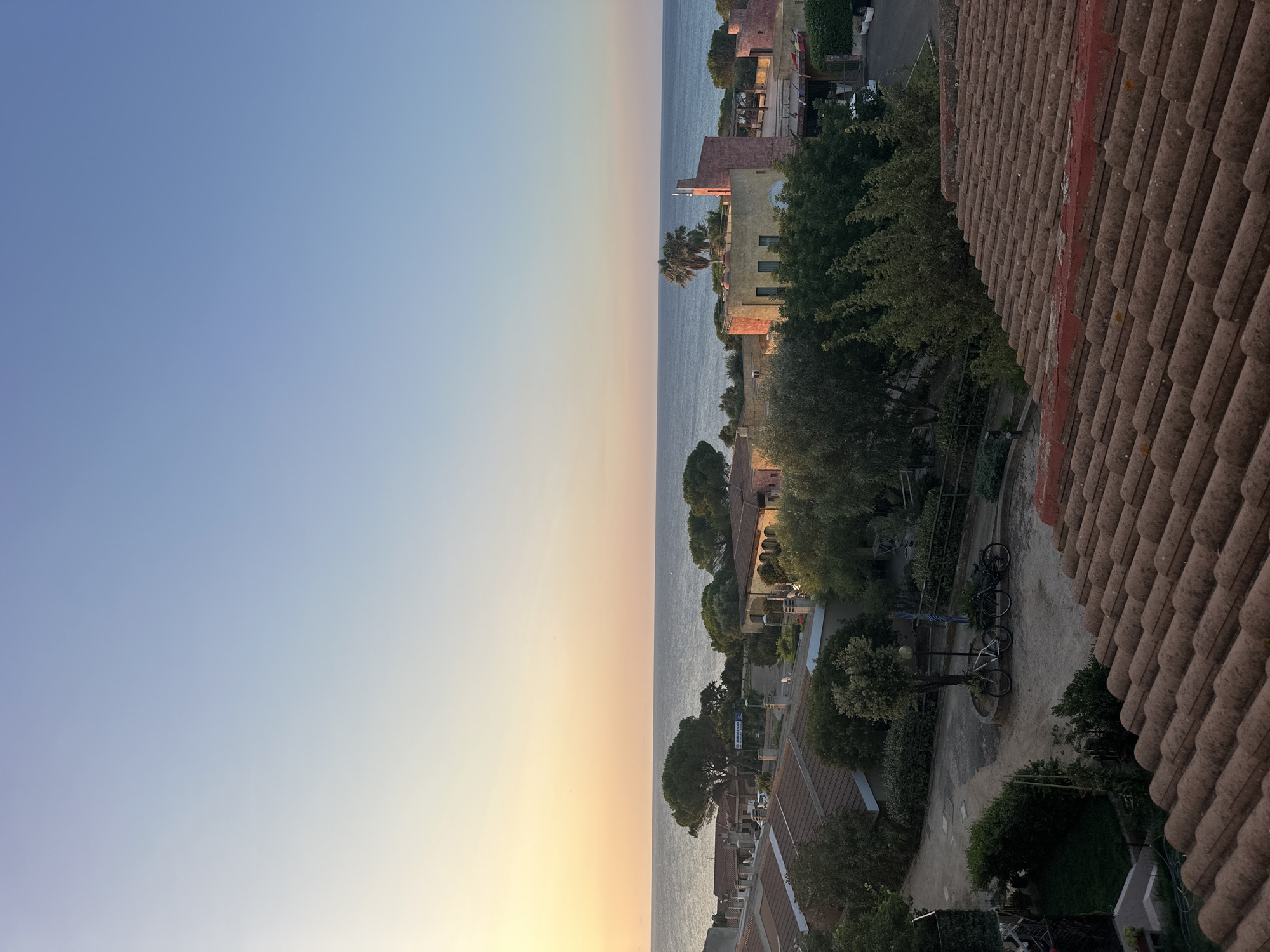 This is a horizontal, eye-level, medium shot of a coastal town at sunset. The sky transitions from a pale yellow-orange at the horizon to a clear, light blue overhead. The sea is visible in the background, calm and reflecting the sky's colors. In the middle ground, a cluster of buildings with terracotta tile roofs and pastel-colored walls are nestled among lush green trees. The architecture appears Mediterranean, with some buildings featuring arches and balconies. A narrow road winds through the settlement, and two bicycles are parked near a lamppost on a gravel path. The foreground is dominated by a close-up view of a tiled roof, its weathered terracotta tiles showing the texture and color variations of age. The scene conveys a peaceful, serene atmosphere, characteristic of a seaside village in the late afternoon or early evening. The location is identified as Biddeputzi/Villaputzu, Italy.