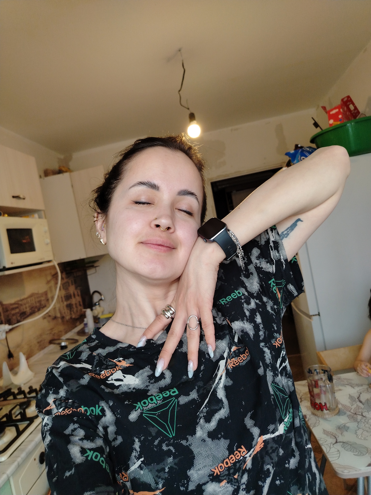 A woman is posing indoors. She has dark hair pulled back, fair skin, and closed eyes. She is wearing a black T-shirt with the "Reebok" logo repeated in various colors and fonts. She has a smartwatch on her left wrist and several rings on her fingers. Her left arm is raised with her hand near her face.
The location appears to be a kitchen in an apartment. In the background, there's a microwave, a stove, and cabinets. There is also a table with a patterned tablecloth and a glass jar on it. Part of a person is visible in the lower right corner of the image at the table.
There is no information to indicate the city location.