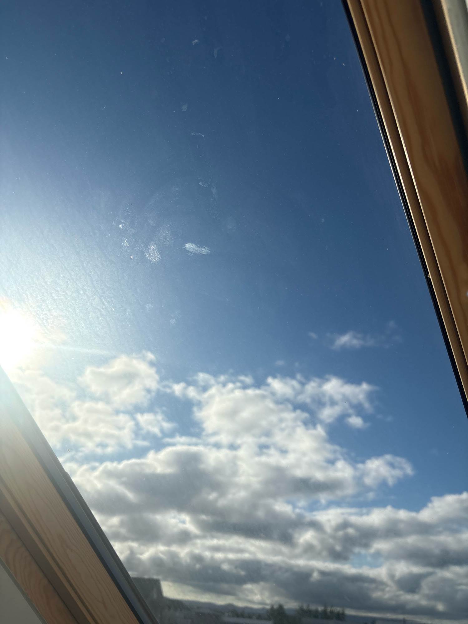 The image depicts a view through a skylight towards a bright blue sky filled with scattered white clouds. The sun is visible on the left side of the frame, casting a bright glare. There are some smudges and streaks on the glass of the skylight. The wooden frame of the skylight is visible along the bottom and right edges of the image. In the distance, below the clouds, a blurry outline of buildings and trees can be seen, suggesting an urban or suburban landscape. The scene is likely a residential or commercial building in Reykjavik, Iceland, given the clear blue sky and the presence of clouds typical of the region. There are no people or animals visible in the image.