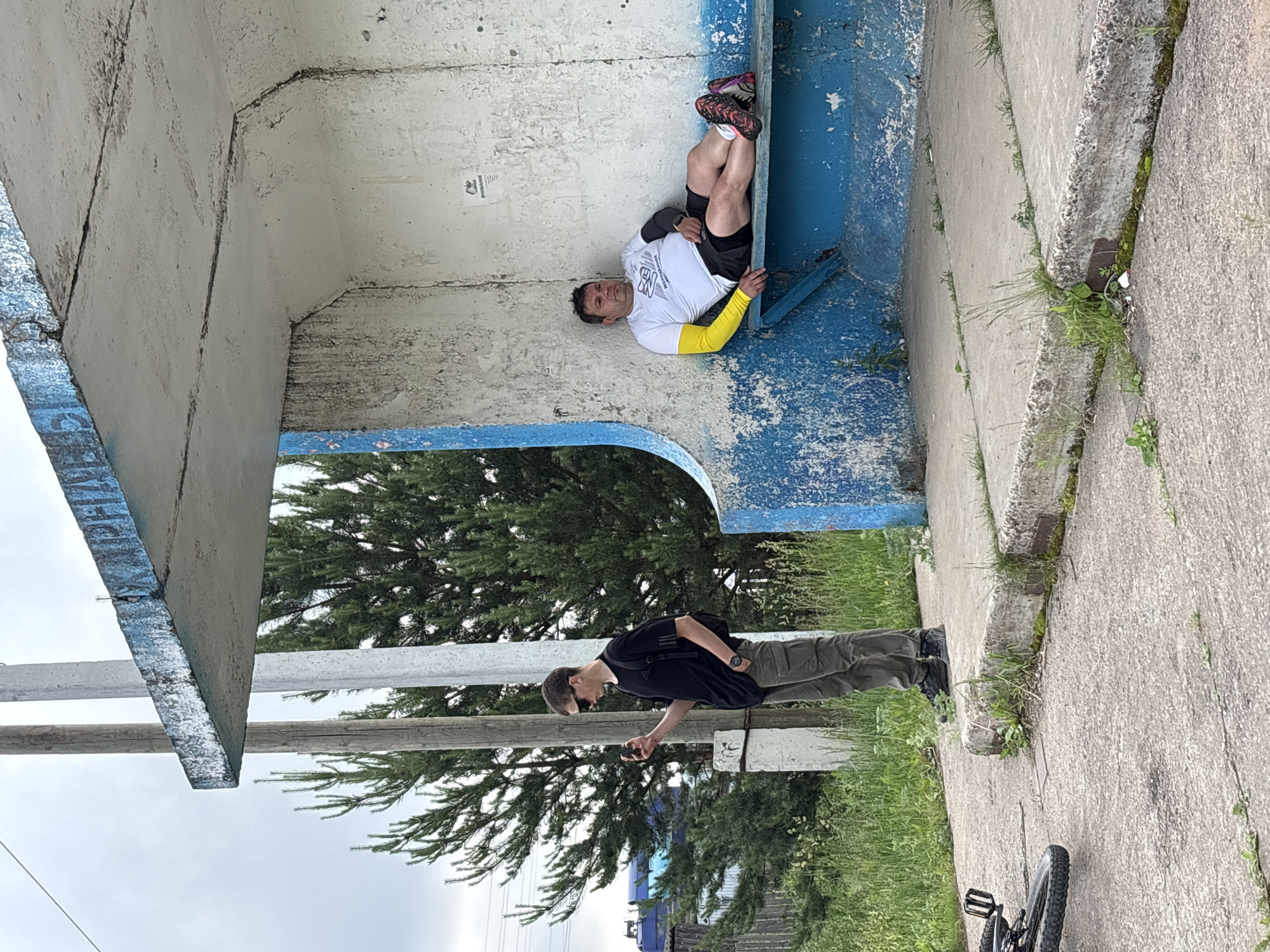 This image depicts a bus stop in Kharnavy, Russia. On the right, a man in a white t-shirt, black shorts, and yellow arm sleeves reclines on a blue bench. He is looking upwards and to his left. On the left, a young man wearing sunglasses, a black t-shirt, and khaki cargo pants stands holding a mobile phone in his right hand. A bicycle is partially visible in the lower left corner of the frame. The bus stop structure is made of concrete and shows signs of wear and tear, with peeling paint on the blue sections. The background consists of green trees and a cloudy sky.