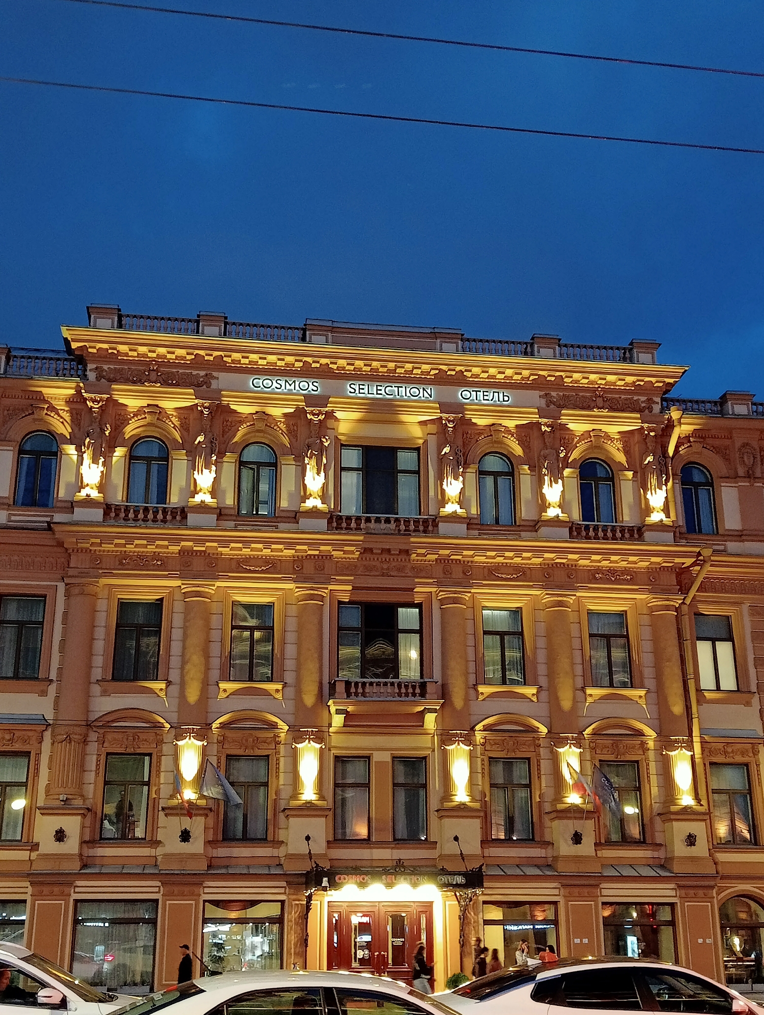 The image captures a detailed exterior view of the Cosmos Selection Hotel in Saint Petersburg, Russia, at dusk. The facade of the building is illuminated by warm lights, highlighting its classical architectural features, including ornate cornices, columns, and statues adorning the upper levels. Multiple arched windows with dark frames are visible across the several stories, some of which reflect the exterior lighting. Two flags are seen hanging from poles between some of the windows on the second floor. The hotel's name, "COSMOS SELECTION" and "ОТЕЛЬ" (Hotel in Russian), is prominently displayed in illuminated lettering across the top of the building. Below the main hotel entrance, large storefront windows display illuminated interiors, suggesting shops or a reception area. Several people are present on the sidewalk in front of the hotel, some walking and others appearing to be in conversation. Two white cars are parked or driving in the foreground, partially obscuring the base of the building and the sidewalk. The sky above is a deep blue, indicating the transition from day to night, with power lines visible horizontally across the upper portion of the frame.