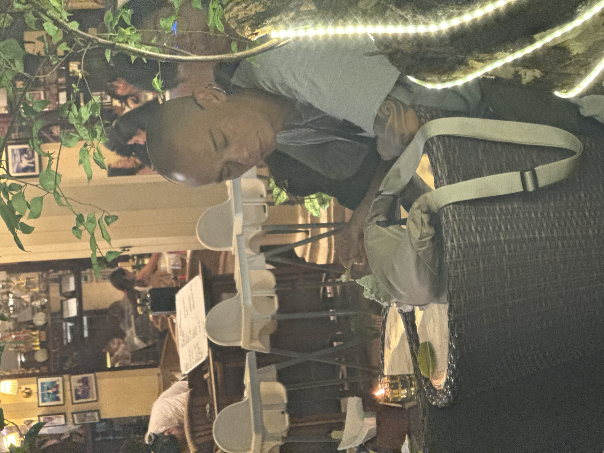 The image depicts an outdoor seating area of a restaurant or cafe, possibly in Denpasar, Indonesia, during evening hours. In the foreground, a bald man with tattoos on his arms is seated at a wicker table, appearing to be smoking or holding a cigarette. A purse or bag is draped over the table. A small candle in a glass holder burns on the table, illuminating the immediate area. In the background, other patrons are visible at tables, and shelves with various items, possibly trophies or decor, are displayed on a wall. High chairs are also set up at some tables. The overall lighting suggests it is nighttime, with artificial lights from lamps and string lights adding to the ambiance.