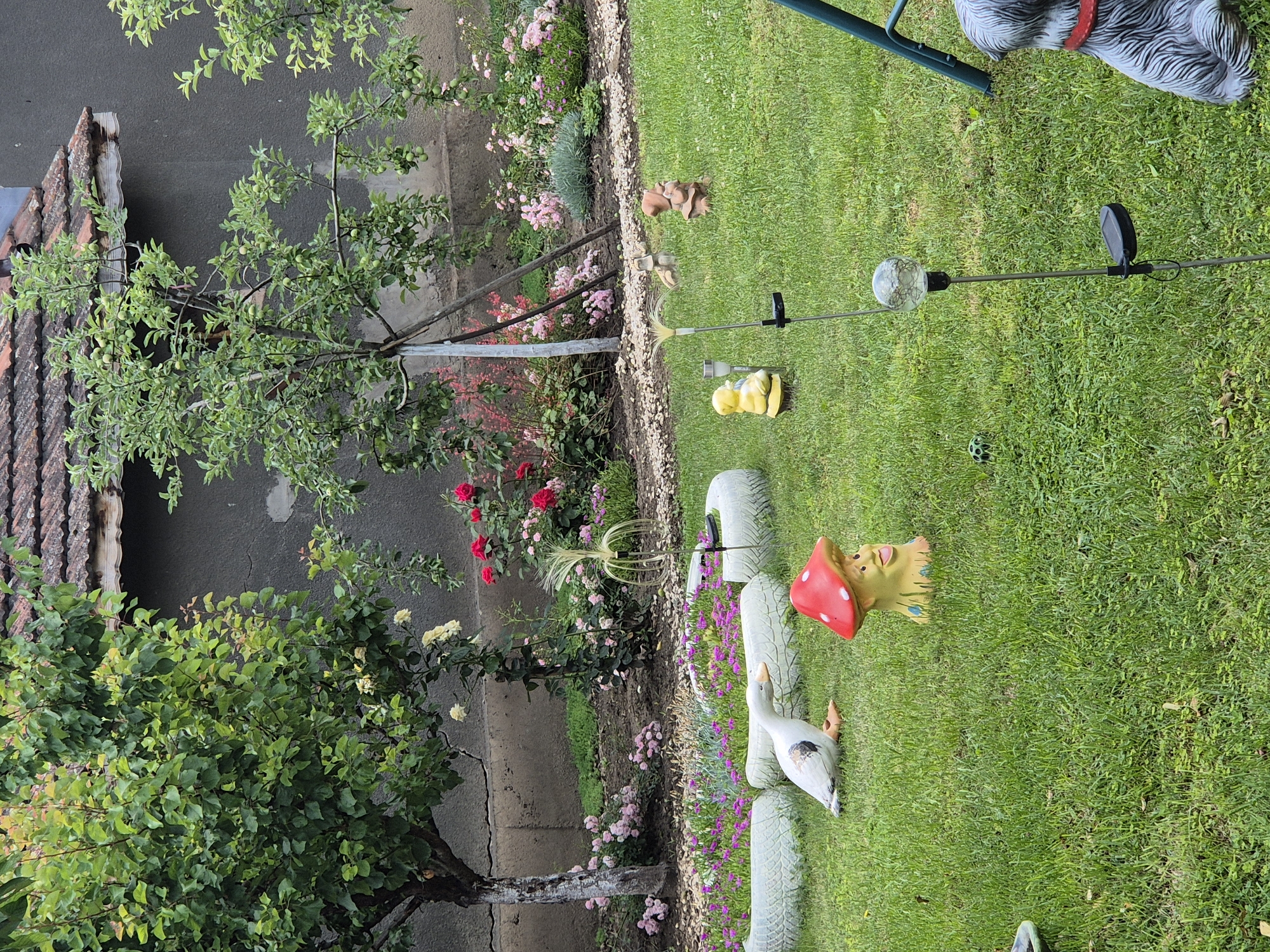 The image depicts a garden scene in an unknown location. The garden features a variety of plants, including rose bushes with red and pink blooms, as well as green trees. Several garden decorations are present, including a white goose, a red-capped mushroom gnome, and a statue of a dog wearing a red collar. Other decorative elements include small animal figurines and a solar-powered garden light. The garden is bordered by a gray wall and a roof with terracotta tiles. The lawn is green and well-maintained.