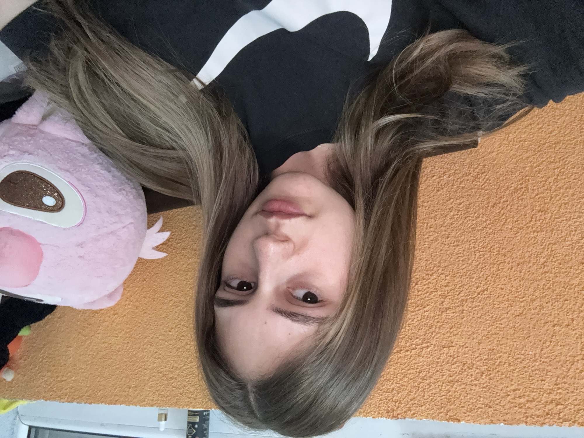 A person is lying down on an orange surface, possibly a floor or bed, with their head near the bottom of the frame. The person has light brown, shoulder-length hair, light skin, and brown eyes. They are wearing a black sweatshirt with a large white logo. To the left of the person's head is a pink, plush toy with a brown eye and white details. In the lower left corner, there is a glimpse of additional toys near a window with a white frame. A bottle is located near the window. There is no indication that this photo was taken in Panagyurishte, Bulgaria.