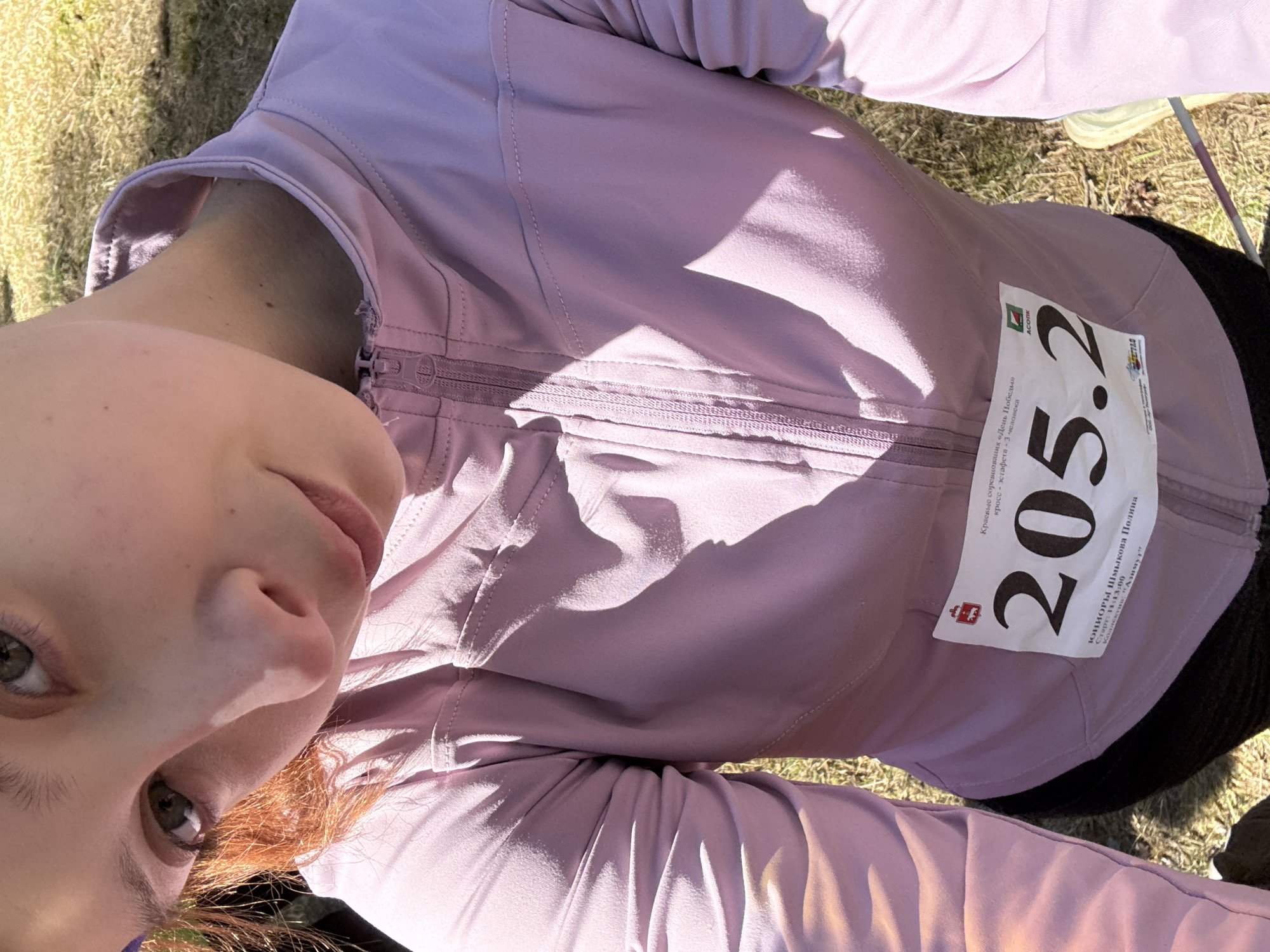 The image shows a person with reddish hair and green eyes, wearing a light purple zippered jacket. Affixed to the right chest of the jacket is a race bib with the number "205.2" in large black numerals. The bib also contains text in Russian, identifying the competition as "Kраевые соревнования «День Победы» кросс-эстафета 3 человека" which translates to "Regional competitions" Victory Day "cross-relay 3 people", indicating a Victory Day cross-country relay race. The text also identifies the participant as "ЮНИОРЫ Шмыкова Полина" which translates to "JUNIORS Shmykova Polina", with their team being "Азимут" meaning Azimuth. The race start time is listed as 11:13:00. The background is out of focus, but shows a grassy area, suggesting an outdoor location in Perm, Russia. The image is taken from a low angle, appearing as though the photographer is looking up at the subject.