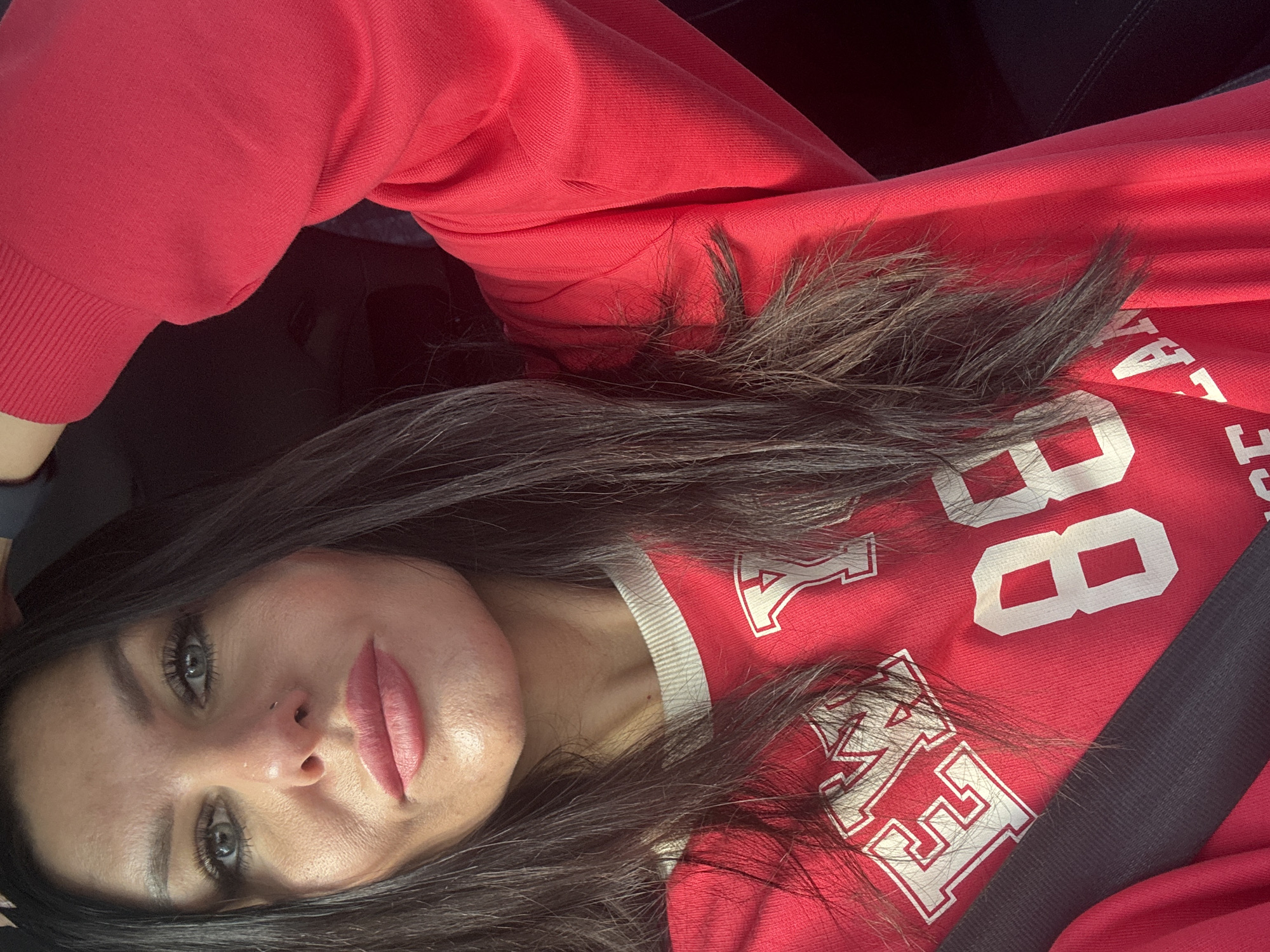 A young woman is lying down in a car seat, wearing a red sweatshirt with white lettering that reads "88" and what appears to be "Marshall" with a collegiate font. Her long, dark hair is spread around her head and on the sweatshirt. She has fair skin, grey eyes, and a nose piercing. She is wearing a seatbelt. The setting is possibly inside a car in Chaikovsky, Russia, but there is no explicit evidence of this in the image.