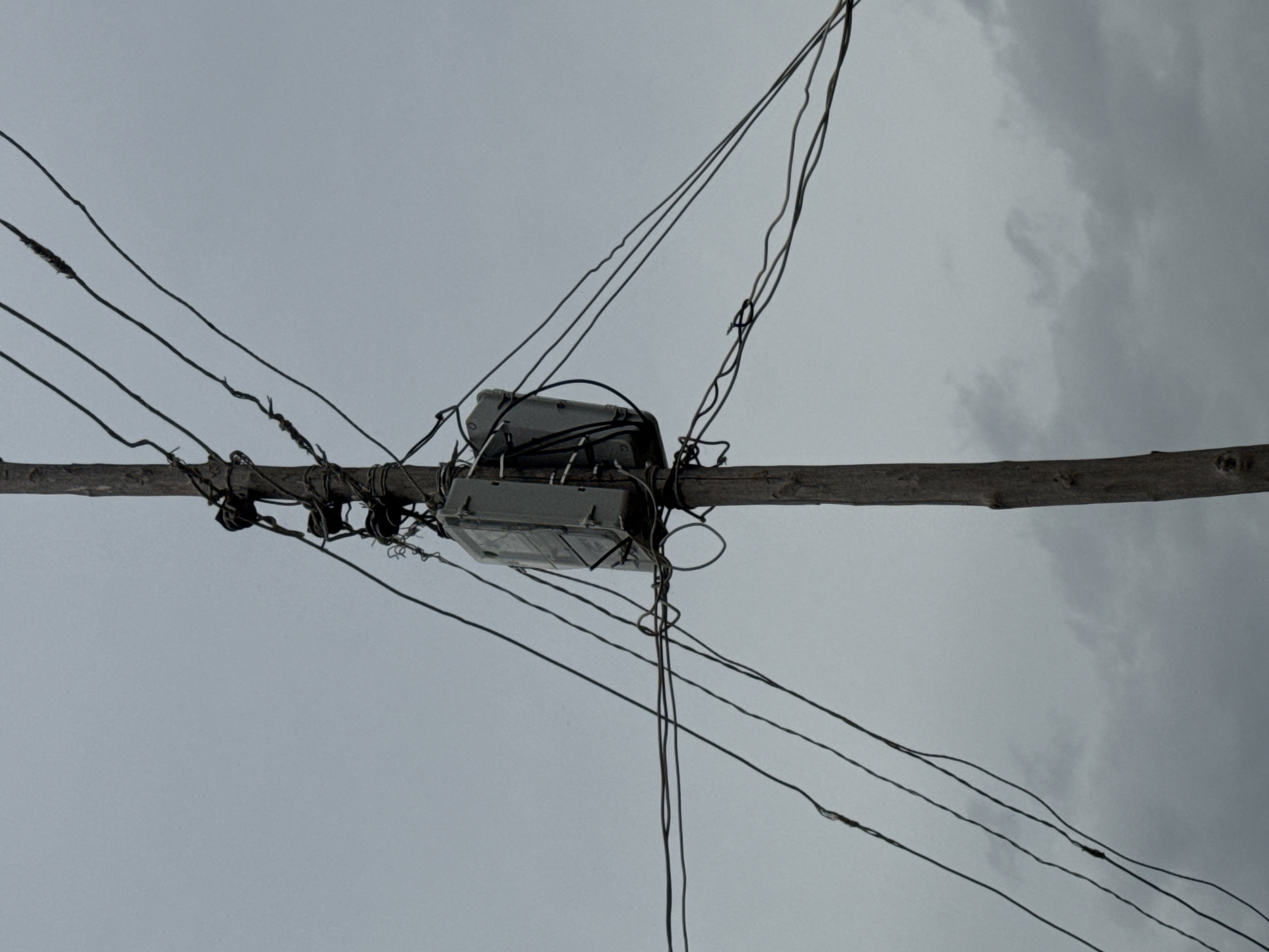 A wooden utility pole stands against a cloudy sky, supporting a gray electrical transformer box and numerous tangled wires. The pole is rough and weathered, with visible knots and a textured surface. The transformer box, rectangular in shape, is attached to the pole by wires and has several smaller wires connected to it. Multiple electrical wires of varying thickness crisscross the frame, attached to insulators on the pole. Some wires appear frayed or damaged. The environment suggests an outdoor setting in Jauro Yinu, Nigeria, likely a residential or commercial area with overhead power lines. There are no people visible in the image, and no specific actions or interactions are depicted, other than the static presence of the utility equipment. The lighting is diffused due to the overcast conditions.