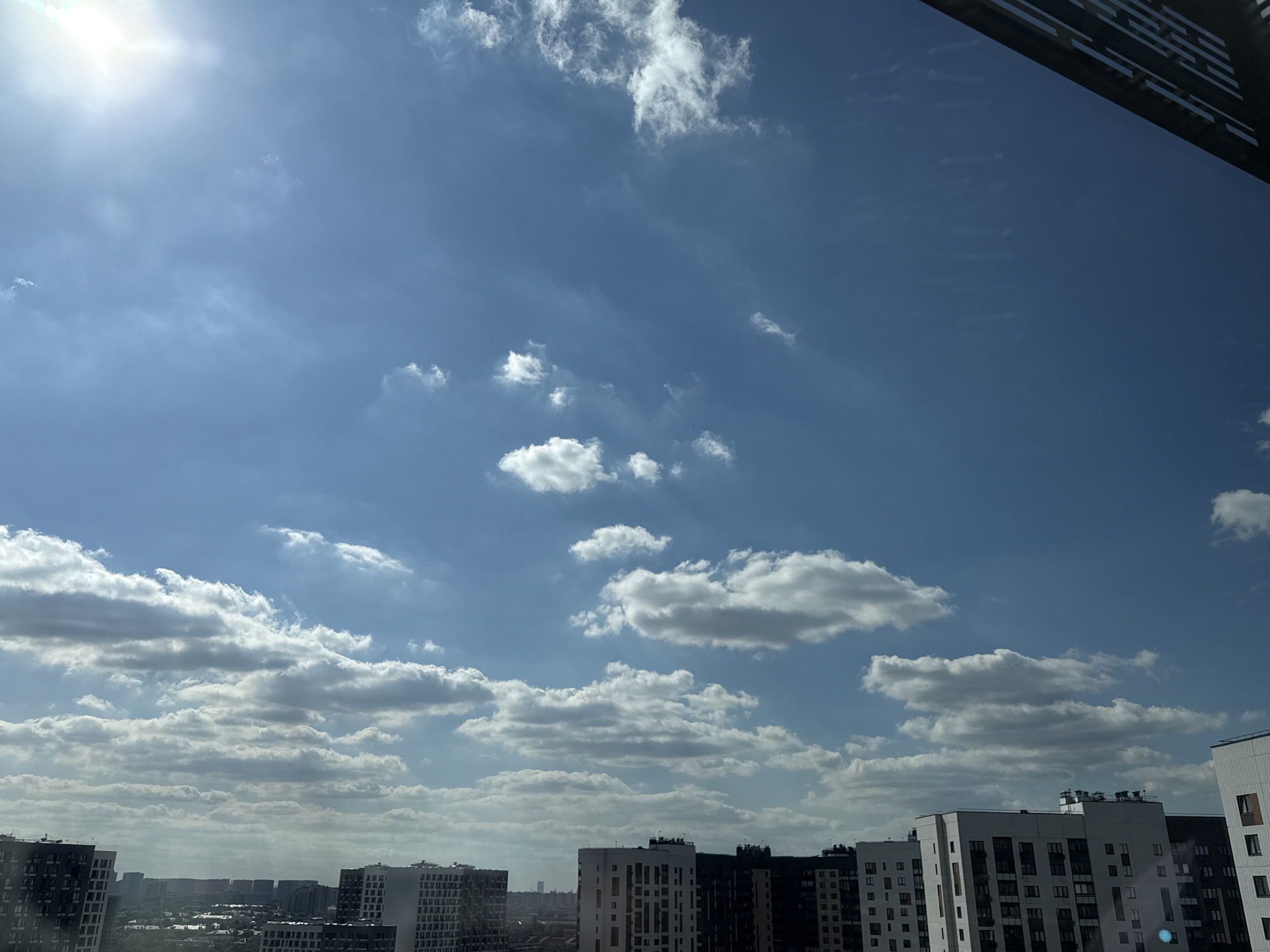 The image depicts a clear sky with scattered white clouds over a city skyline. The sun is bright and visible in the upper left corner of the frame, casting a warm glow. The sky is a vibrant shade of blue, with wispy cirrus clouds appearing higher up and more clustered cumulus clouds closer to the horizon.
In the foreground and mid-ground, a dense urban landscape is visible. Numerous apartment buildings of varying heights and architectural styles dominate the scene. Some buildings are modern with clean lines and light-colored facades, while others appear older and darker. The buildings are densely packed, suggesting a residential or mixed-use urban environment. In the far distance, a hazy view of more buildings and possibly some greenery can be seen, indicating the extent of the city.
The overall mood is one of a bright, sunny day in a populated urban area. The abundance of clouds suggests fair weather, perhaps with a slight breeze. The composition, with the sky taking up a significant portion of the frame, emphasizes the vastness of the sky and the daylight. There is no visible text in the image. The setting appears to be Lyubertsy, Russia, based on the provided location context. The time of day is clearly daytime, likely late morning or early afternoon, given the sun's position and intensity.