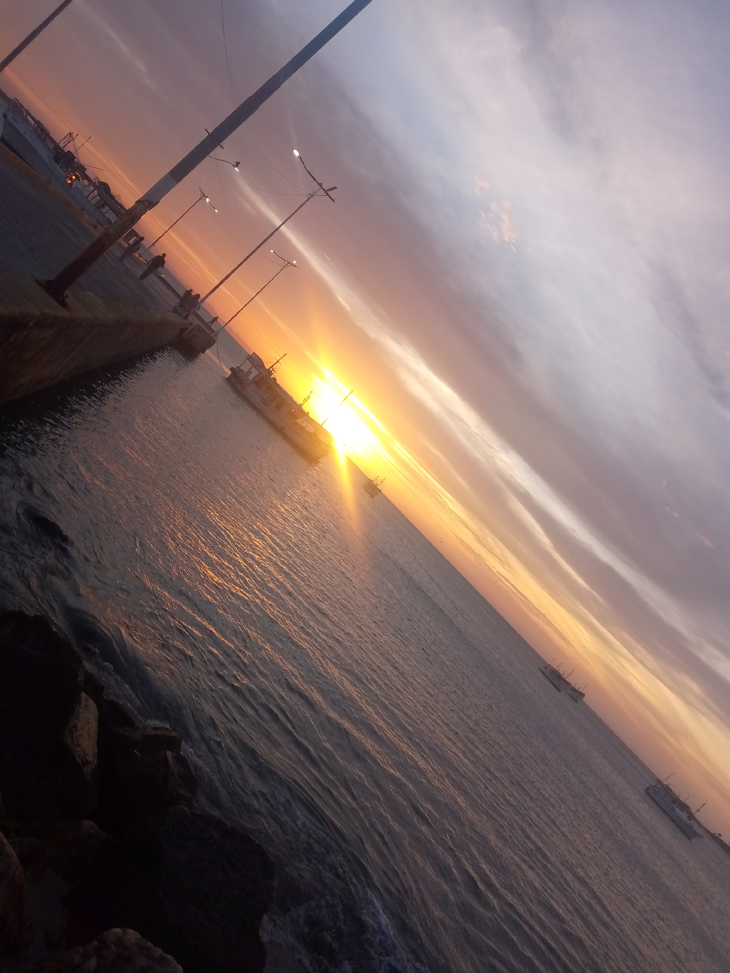 The image captures a sunset scene at a waterfront in an unknown city. The sun is setting over the water, casting a golden glow across the scene. A boat is positioned in the water near the shore, partially silhouetted against the sun's glare. On the shore, there is a paved area with streetlights and what appears to be a low wall or barrier separating it from the water. There are also some people on the shore, possibly walking or standing along the waterfront. The sky is a mix of colors, with orange and yellow near the horizon fading into purple and gray higher up. Rocks or other structural elements are visible in the foreground on the left side of the frame. In the distance, other boats can be seen further out in the water.