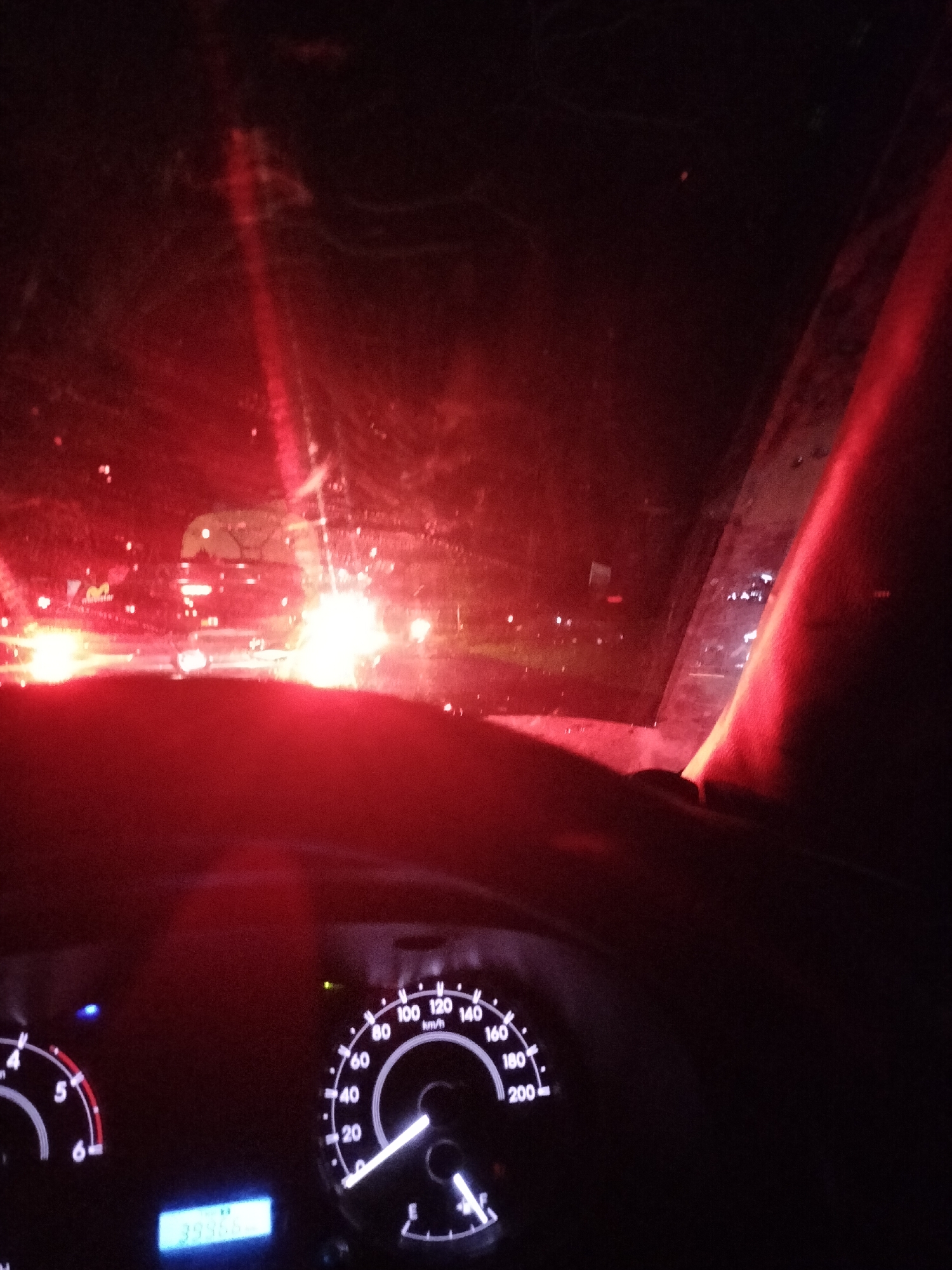 The image is a nighttime, interior shot from the driver's perspective inside a vehicle, likely in Alue Dua Bakaran Batee, Indonesia. Red light from outside, possibly from streetlights or other vehicles, reflects intensely on the windshield, obscuring much of the view. A portion of the dashboard is visible, including a speedometer with markings up to 200 km/h and an odometer displaying the number "39966". The bright red glare on the windshield creates a hazy, atmospheric effect. The overall scene suggests the vehicle is either stopped or moving slowly in traffic.
