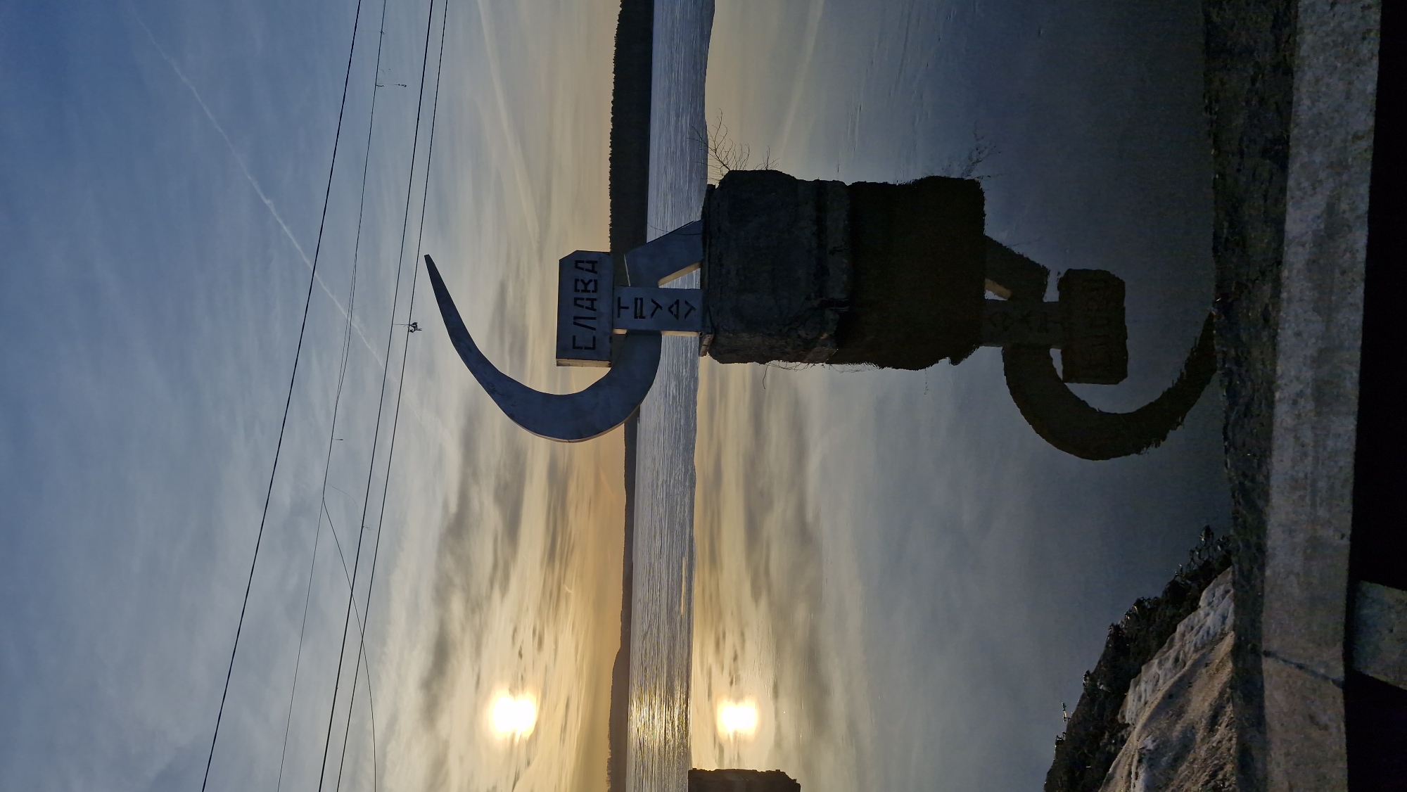 The image, correctly oriented, displays a serene sunset over a wide body of water in Otchor, Russia. In the foreground, on the right, stands a large, metallic, crescent-shaped monument resembling a sickle, rooted in a rough stone base. Attached to this base is a rectangular sign with the Cyrillic word "СЛАВА" (SLAVA), meaning "GLORY," written horizontally. Below "СЛАВА," two stylized symbols are visible: one appears as an inverted capital "T" and the other as a right-pointing chevron or arrow.
The sun, low on the horizon, casts a warm orange and yellow glow, creating two distinct bright reflections on the calm water. The water extends into the distance, meeting a dark, low landmass or shoreline on the far horizon. The sky is a gradient of blue overhead, transitioning to soft yellow and orange near the horizon, dotted with wispy clouds and a faint, thin contrail. Multiple power lines stretch across the upper portion of the frame. The monument's reflection is clearly visible in the water below, and a concrete or stone embankment forms the immediate foreground on the bottom right. The scene conveys a peaceful and contemplative atmosphere.