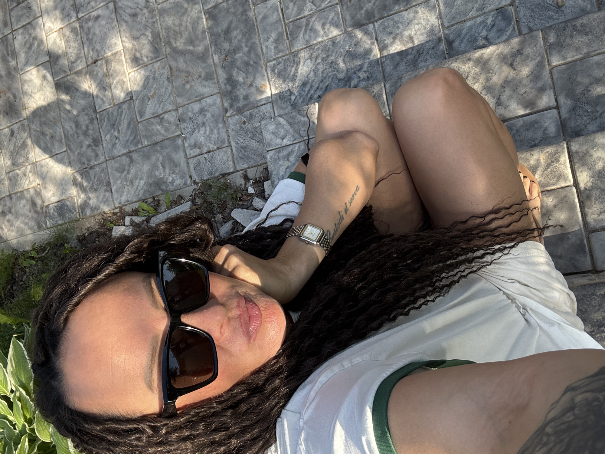 A single individual is depicted in an outdoor setting within the city of Chaikovsky, Russia. The person, appearing to be an adult female, is positioned in a low squat or kneel on grey, rectangular pavers. She is wearing a white t-shirt with green trim on the sleeves, dark rectangular sunglasses, and a square-faced metallic watch on her left wrist. Her dark, dreadlocked or heavily textured hair falls over her shoulders and chest. A tattoo with script is visible on her left forearm. Her right hand is placed against her face, near her chin, with her lips slightly pursed. Strong sunlight from the upper right illuminates her and casts a distinct shadow of her upper body onto the pavers to her right. Green foliage, resembling hostas, is visible in the upper left background, bordering the paved area.