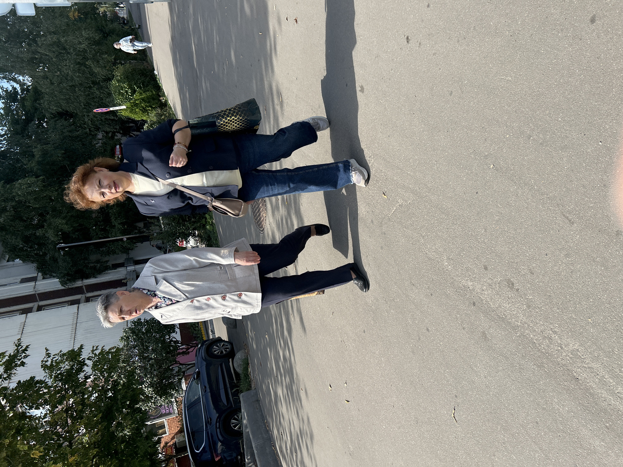 Two women walk on a paved street in Moscow, Russia. The woman on the left wears a light-colored jacket, dark pants, and black shoes. She has short, gray hair and is looking to her left. The woman on the right wears a dark blazer over a light-colored shirt, blue jeans, and sneakers. She has auburn hair and is looking forward. A dark SUV is parked on the left side of the street. Trees and a building are visible in the background. The sun casts shadows on the ground.