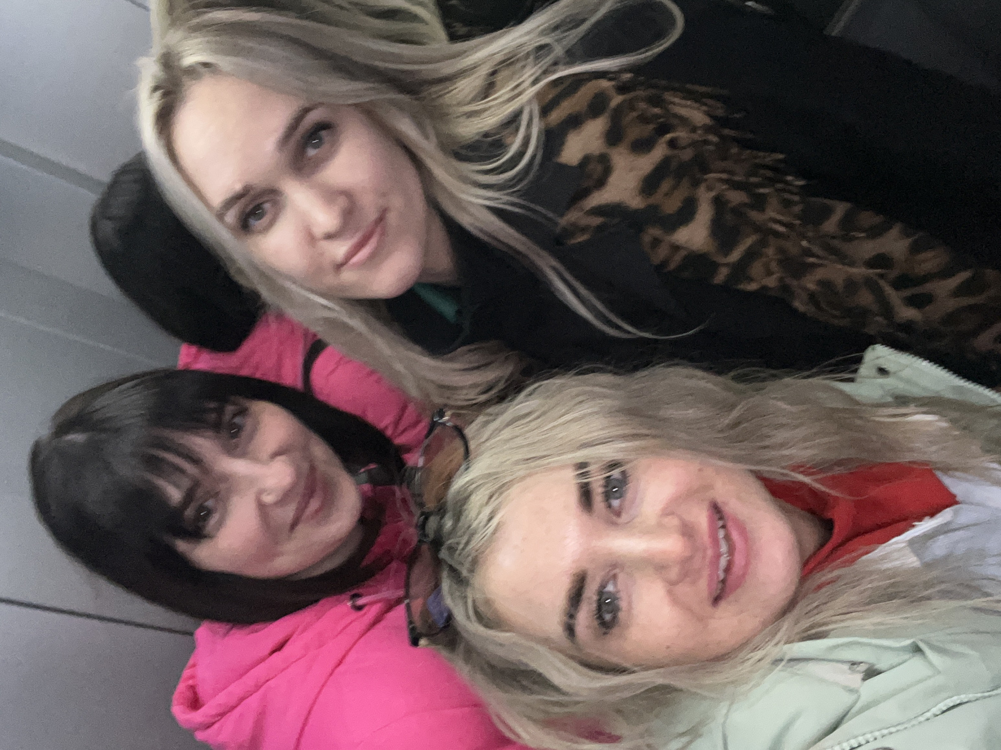 Three women are posing for a selfie. One woman is wearing a pink jacket and has dark hair. Another woman is wearing a black coat with an animal print collar. The third woman has blonde hair and is wearing a light colored jacket. They are possibly in a car or bus. The location is unknown.