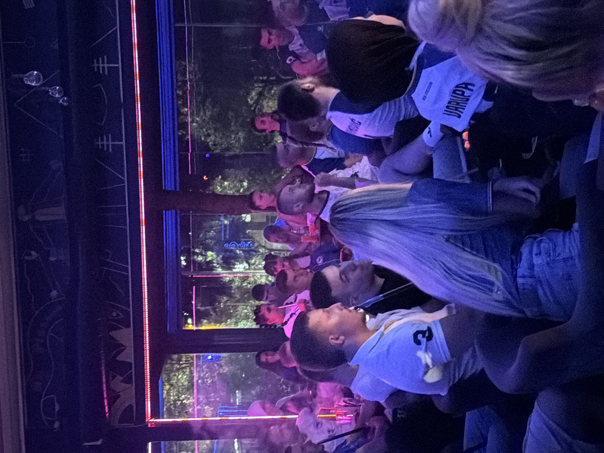 The image depicts a group of people gathered in what appears to be an indoor smoking lounge or cafe, possibly in Travnik, Bosnia and Herzegovina, given the context. The atmosphere is dimly lit with vibrant neon and LED lighting, primarily in shades of pink and blue, casting a colorful glow on the patrons and the surroundings. Many individuals are seated, some appearing to be smoking shisha pipes, indicated by the presence of hoses and smoke. The people are casually dressed, and some are wearing sports-like jerseys, one of which has the name "JARUPA" visible on the back. The environment includes seating arrangements, tables with items on them, and decorative elements on the walls and ceiling. The overall scene suggests a social gathering in a relaxed, potentially entertainment-oriented venue.