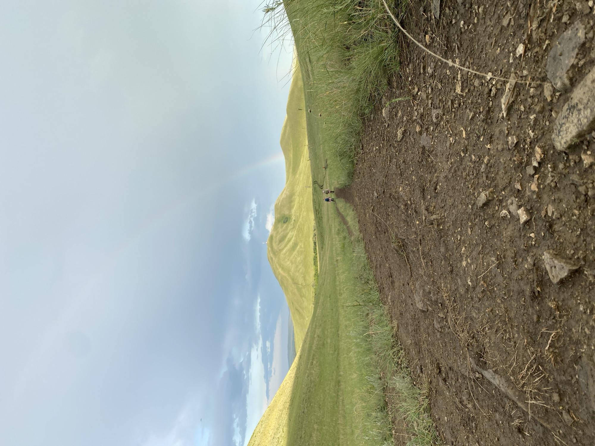 In the city of Verkhneozernoye, Russia, a dirt path winds through rolling green hills under a cloudy sky. A faint rainbow arcs across the horizon, suggesting recent rainfall. Several figures are visible walking along the path in the distance, heading towards a prominent, rounded hill topped with a structure. The foreground of the path is muddy and strewn with small rocks and dry vegetation. The grassy slopes on either side of the path are tall and appear somewhat unkempt.