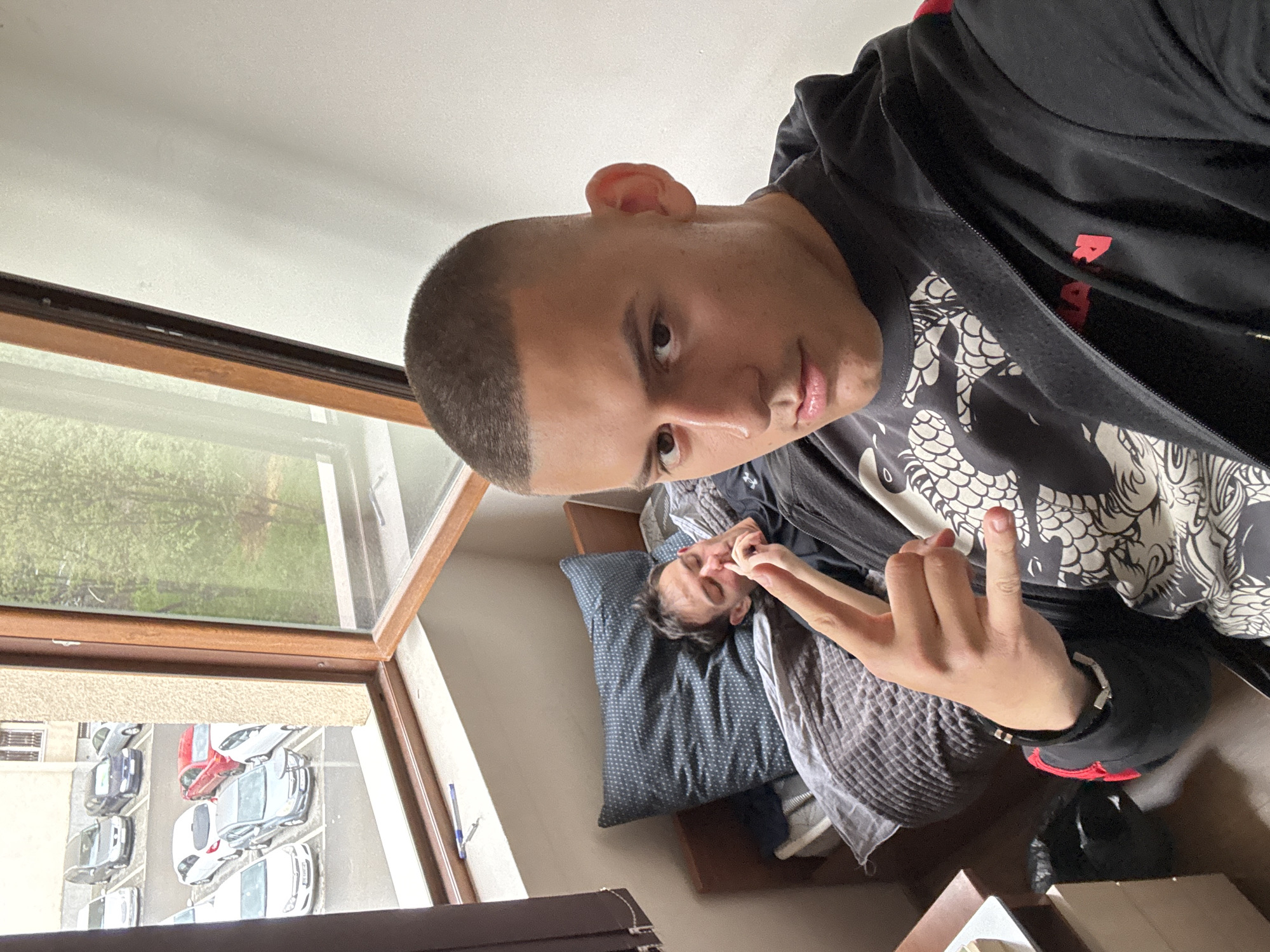 The image is a vertical selfie taken indoors, capturing two individuals and a view outside a window. In the foreground, a young man with a buzz cut is visible, looking directly at the camera with a slight smirk. He wears a black zip-up hoodie over a t-shirt featuring an intricate white dragon-like graphic. His right hand is raised, making a gesture resembling a "rock on" sign. A red detail, possibly with partial text "RAPH" or "RALPH", is visible inside his hoodie zipper.
In the mid-ground, another person is lying in bed, partially obscured. They are resting on a dark blue pillow with white dots and appear to be covering their face or rubbing their nose with their hands. Grey bedding covers them.
To the left, a large, open window with a brown wooden frame reveals the exterior. The upper portion of the window shows lush green foliage, suggesting trees or bushes. Below, a parking lot is visible, with several cars (white, red, grey) neatly parked in rows, indicative of a residential street or complex. A pen rests on the window sill.
The bright natural light suggests it is daytime. The overall scene conveys a casual, indoor moment, possibly in an apartment or residence in an urban area, consistent with Plovdiv, Bulgaria. The man in the foreground appears confident and playful, while the person in the background seems relaxed.