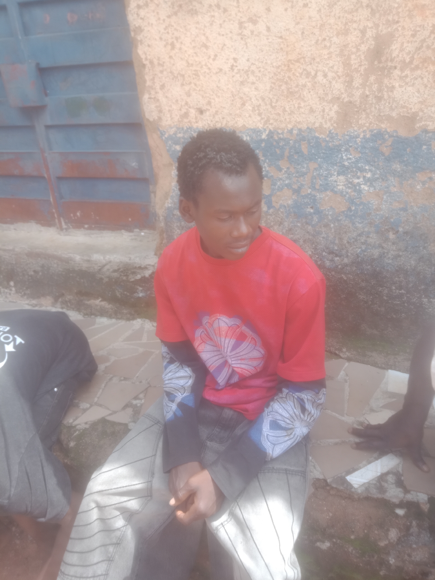 A medium shot shows two young Black males sitting on a paved area with a patterned stone surface. The male in the foreground is wearing a red long-sleeved shirt with a circular, floral-like graphic on the chest and a black long-sleeved undershirt. His pants are light gray with vertical pinstripes. He is looking down and to the right. The second male, to the left and partially obscured, is wearing a black t-shirt with a graphic design on the left shoulder. His pants are dark gray. The background features a rough, textured wall, painted in patches of beige, blue, and rust, with a corrugated metal gate on the left. The lighting is bright, casting shadows.