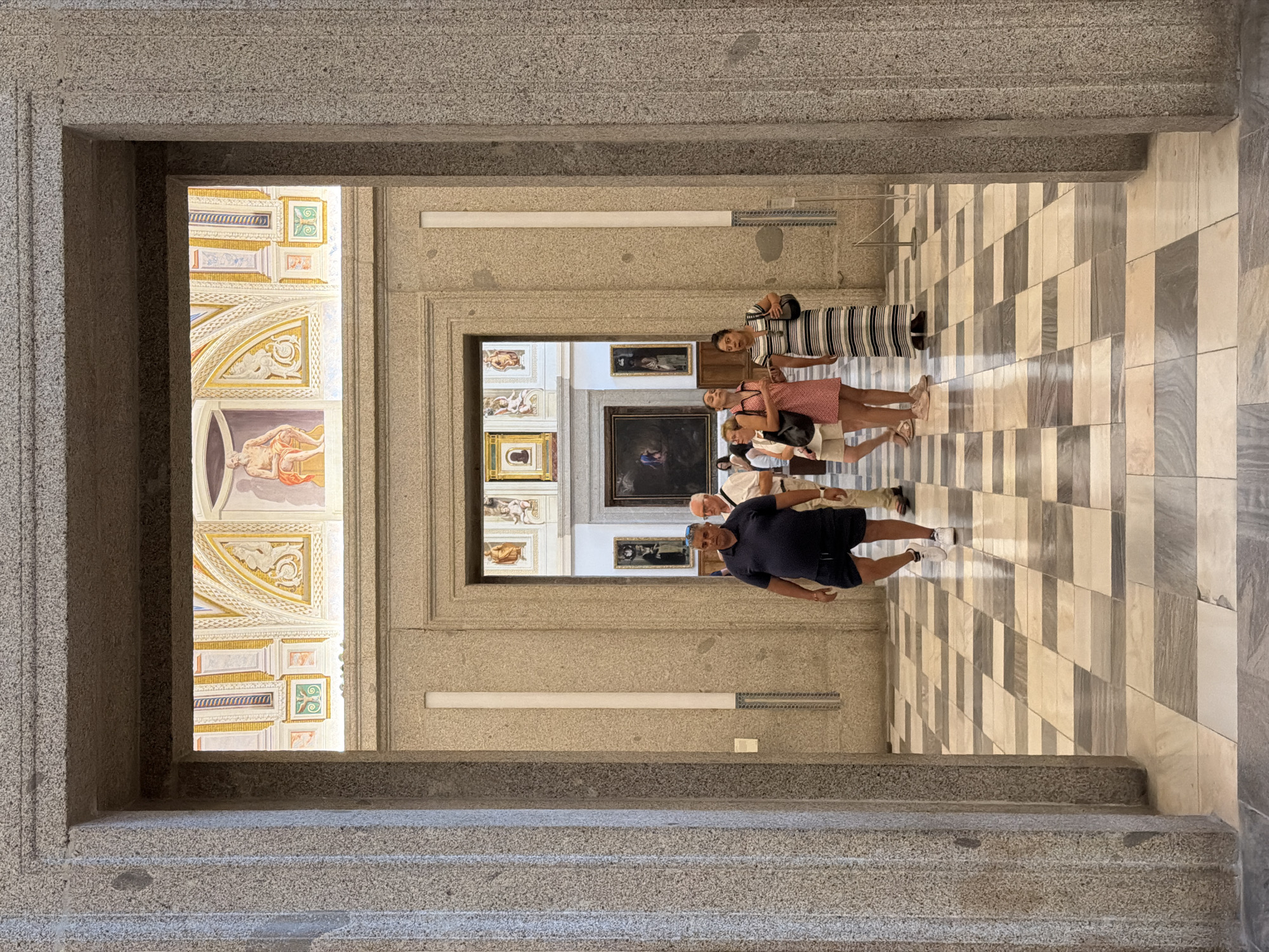 In San Lorenzo De El Escorial, Spain, within a grand hallway of what appears to be a historical building or museum, the image captures a view through a large stone archway. The hallway features a black and white checkered marble floor that creates a visually striking pattern. Several individuals are walking through the hallway, some towards the viewer and others away.
In the foreground, to the right and left, are large, textured stone columns framing the entrance. Beyond the archway, the hallway opens up to reveal more of the building's interior. Prominently displayed on the walls are various framed artworks, including paintings and decorative murals, suggesting an art gallery or a historically significant location. The ceiling area above the hallway is adorned with intricate murals featuring figures and architectural elements, illuminated by soft lighting.
The people visible in the scene are casually dressed, indicating they are likely visitors. One man in the foreground is wearing a dark blue polo shirt and dark shorts, walking towards the camera. To his right, a man in a white shirt and beige pants is also walking in the same direction. Further into the hallway, a group of three women is walking away from the camera. One woman in the center wears a pink dress, and another to her right is in a black and white striped dress. The overall atmosphere suggests a leisurely visit to a place of cultural and artistic significance.