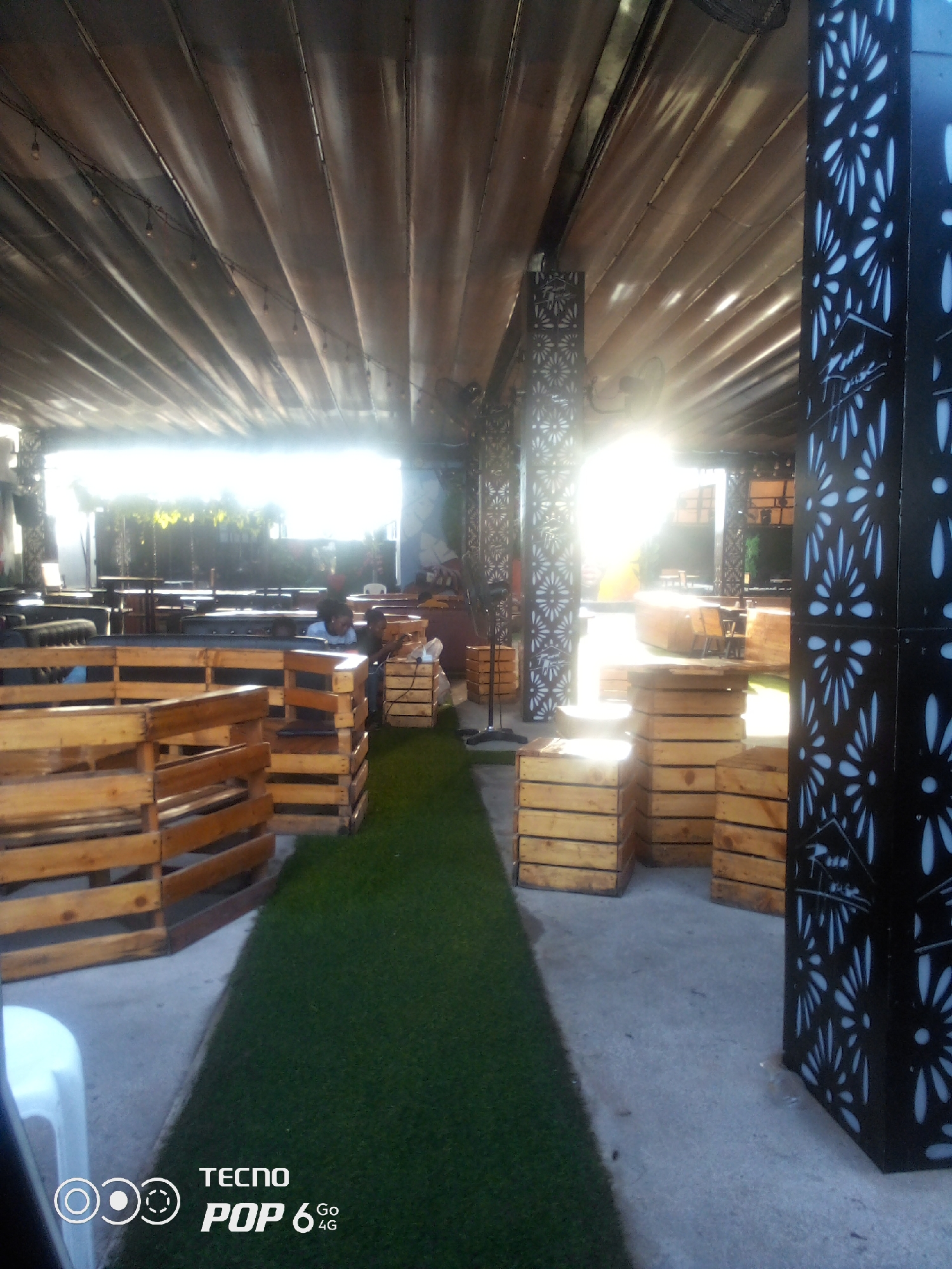 The image depicts an outdoor seating area, possibly a bar or restaurant, in the city of Howayi, Nigeria. The space is covered by a corrugated metal roof, supported by decorative pillars with a white floral pattern against a black background. In the foreground, there are wooden pallet sofas and stacked wooden crates, serving as furniture and dividers. Artificial turf covers the ground in some areas, creating a contrasting green surface.
Several tables and chairs are visible in the background, arranged in rows. Sunlight streams in from an open area, creating bright highlights and casting shadows. Two individuals are seated at a table in the middle ground, appearing to be engaged in conversation or simply relaxing. One person is wearing a blue shirt, and the other is partially obscured. A white plastic chair is visible in the lower left corner of the frame. The overall impression is one of a casual, outdoor gathering space. The Tecno Pop 6 4G logo is watermarked in the bottom right corner.