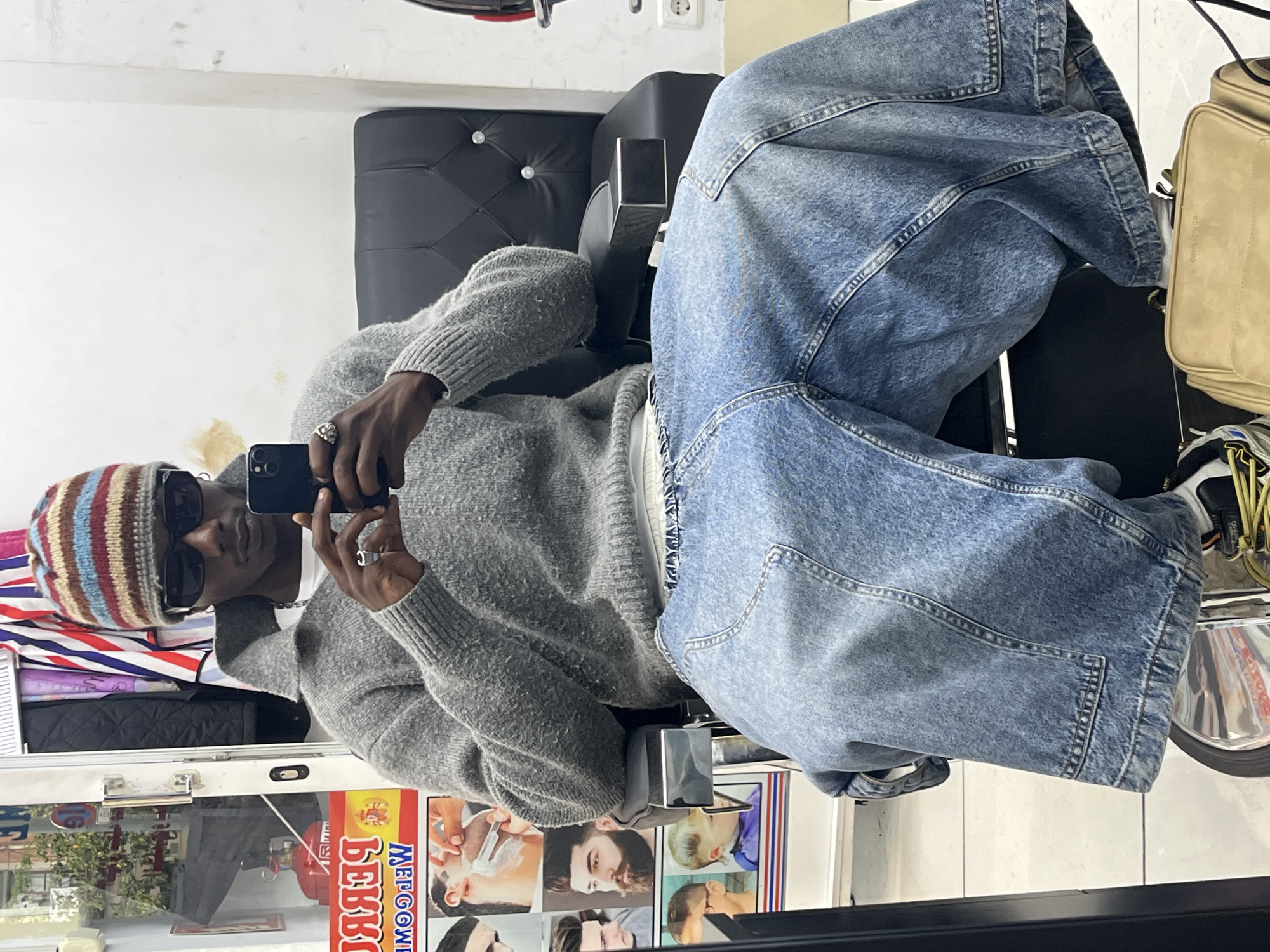 The image depicts a person in a barber's chair taking a selfie using a smartphone. The individual is wearing a striped knit hat, sunglasses, a light grey knit sweater, and light wash denim pants. The person's headwear features horizontal stripes in hues of blue, red, brown, and beige. The barber's chair has black upholstery and is surrounded by a mirrored surface. A beige-colored shoulder bag and a pair of athletic shoes are visible near the chair. The mirrored surface showcases a storefront advertising "Бевоое" in Cyrillic script, suggesting the presence of a barber shop. A barber shop advertisement featuring photos of men undergoing grooming services is also seen. The environment reflects the interior of the barber shop, with various items and equipment visible. Given the Cyrillic script on the storefront, it is unlikely this scene is set in Barcelona, Spain. It is more likely located in a region with a strong Russian-speaking population.