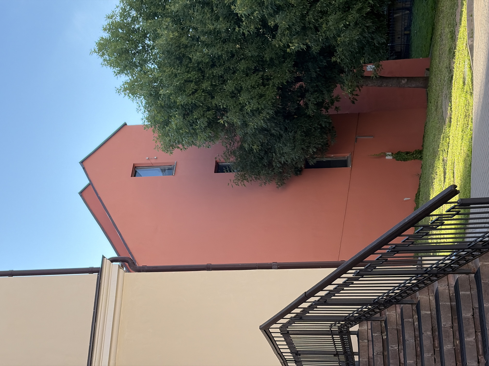 The image captures a view of modern residential buildings and an outdoor staircase with a metal railing. To the left, a building with a light beige facade features a prominent cornice and a dark downspout. Adjacent to it, a building with a vibrant coral-red facade has a steeply pitched roof and several windows. A large, leafy green tree is positioned between these buildings and a dark metal fence in the background. The scene is set against a clear blue sky, with a well-maintained lawn and paved walkways. Based on available data, this location is identified as being in Minsk, Belarus, specifically at the intersection of Pobediteley Avenue and Zhuravleva Street. No people are visible in the image.