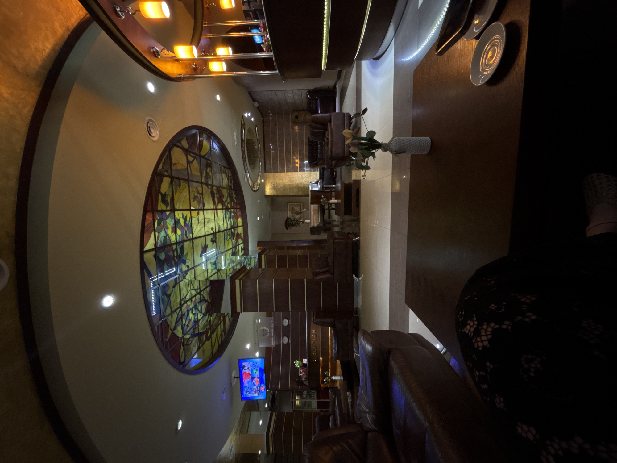 The image depicts the interior of a hotel lobby, likely in Yerevan, Armenia, based on the presence of the "ARARAT HOTEL" sign on the reception desk. The scene is dimly lit, with warm, ambient lighting emanating from overhead fixtures and accent lighting.
In the foreground, a dark wooden coffee table is visible, with a white patterned vase containing a small bouquet of yellow flowers placed on it. To the right of the vase, a white saucer with a swirl design is partially visible. A portion of a dark brown leather couch is in the lower left of the frame, and another dark leather armchair is situated behind it.
The mid-ground features a reception desk with a dark wood finish, labeled "RECEPTION" and displaying the "ARARAT HOTEL" signage. Behind the desk, a large television screen is mounted on the wall, displaying a colorful image. Several cylindrical, pendant light fixtures with warm yellow illumination hang from the ceiling above the reception area.
The ceiling is a prominent feature, with a large, circular stained-glass panel depicting a floral or vine motif in vibrant yellow, green, and red hues. This stained-glass element is illuminated from within, creating a striking visual. Other ceiling elements include recessed lighting and a dark, curved trim.
In the background, more seating areas with dark leather furniture can be seen, along with tall, dark wooden pillars that support the ceiling. A large, golden-colored wall panel adds a touch of opulence to the space. The floor is a polished tile, reflecting the ambient light. The overall atmosphere suggests a luxurious and inviting hotel lobby.