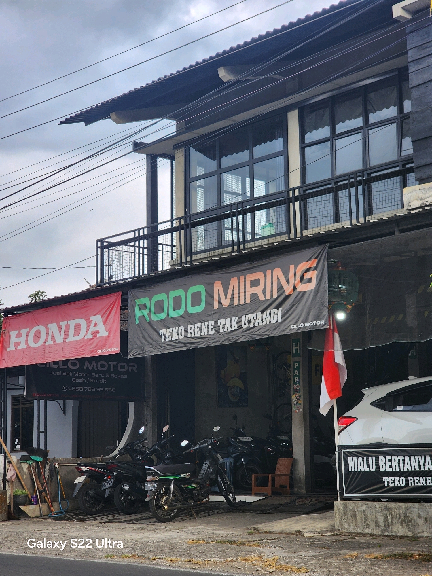 This is a medium shot photograph taken outdoors on a cloudy day, likely in Salaman, Indonesia. The image features the facade of a two-story building with a black wooden exterior on the upper level and a black metal railing on a balcony. The lower level appears to be a shop or garage, with motorcycles parked outside. A red banner with the white "HONDA" logo is hung on the left side, and a black banner with green "RODO MIRING" text is hung in the center. Below the banners, a black banner reads "CILLO MOTOR Jual Beli Motor Baru & Bekas Cash / Kredit" and a phone number is visible.
In front of the building, several motorcycles are parked. To the left, a collection of brooms and cleaning tools are propped against the wall. A white car is parked on the right side of the image, in front of a black banner with white text that says "MALU BERTANYA TEKO RENE". An Indonesian flag, red and white, is attached to a pole above the car and banner. The ground in front of the building is paved and shows signs of wear. The overall impression is of a commercial establishment, likely a motorcycle dealership or repair shop.