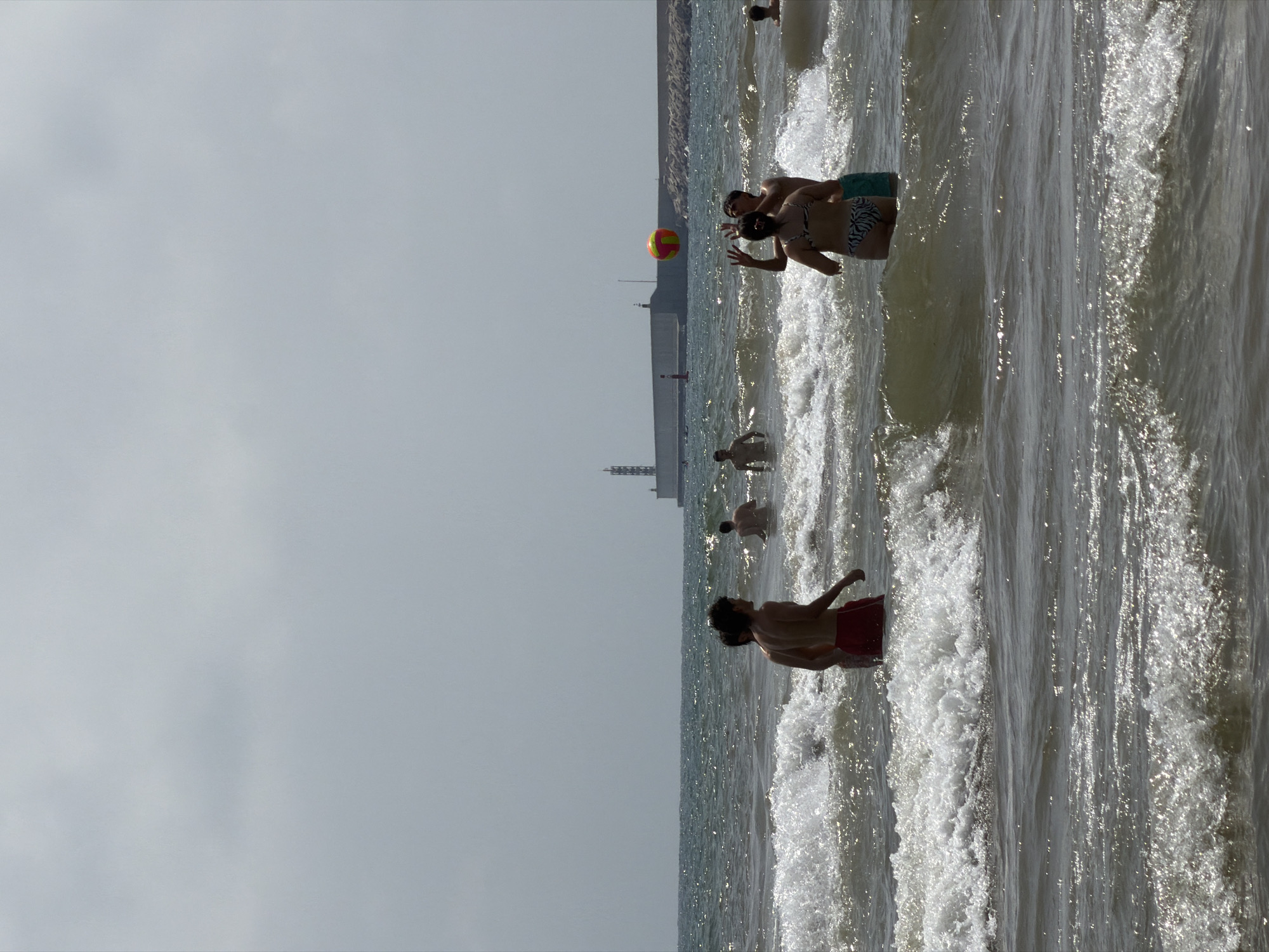 The image depicts a group of people in the ocean, playing with a ball. The water is choppy with visible waves. In the background, a long pier or breakwater extends into the water, with a lighthouse-like structure at its end. The sky is overcast. The people appear to be enjoying recreational activities on a sunny day.