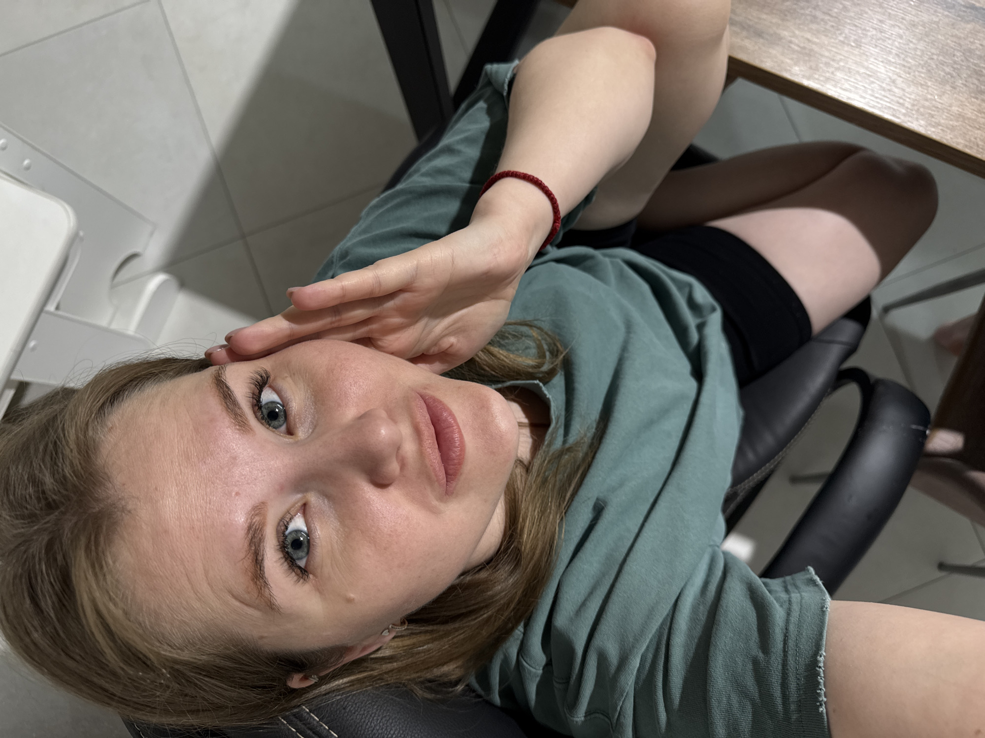 A young woman with light brown hair and blue eyes is the central subject of the image, depicted from a high-angle perspective, possibly a selfie. She is reclining or sitting in a black office chair, looking directly towards the camera with a neutral expression. Her right arm is bent with her hand resting near her forehead, while her left arm is also bent, obscured slightly by her body. She wears a relaxed-fit, seafoam green or muted teal t-shirt or hoodie and black shorts. A thin red string bracelet is visible on her left wrist.
The setting appears to be an indoor space, possibly a room in a home or office, with a light-colored tiled floor that shows distinct shadows, indicating a strong light source. Part of a wooden table or desk is visible in the upper right corner of the frame. White or light-colored furniture or structures are partially seen on the left side of the image. The lighting casts clear, defined shadows on the floor and objects, suggesting artificial light or directional natural light from an unseen window. There is no visible text in the image. The overall impression is one of a casual, relaxed moment in an indoor environment in Gornyi (Dvurechenskoe S/P), Russia.