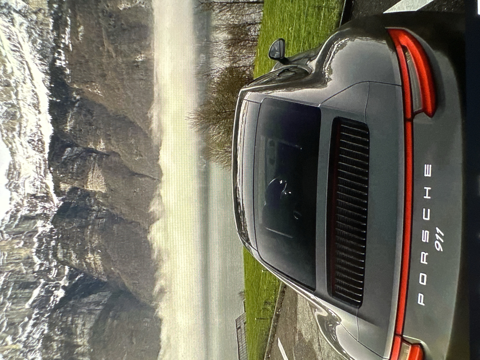 The image shows the rear of a gray Porsche 911 parked in front of a landscape featuring a body of water and mountains. The vehicle is parked on a paved surface next to a grassy area. The background suggests a lakeside or coastal mountain setting, with some evidence of mist or fog obscuring the distant shore. The location is not Yerevan, Armenia.