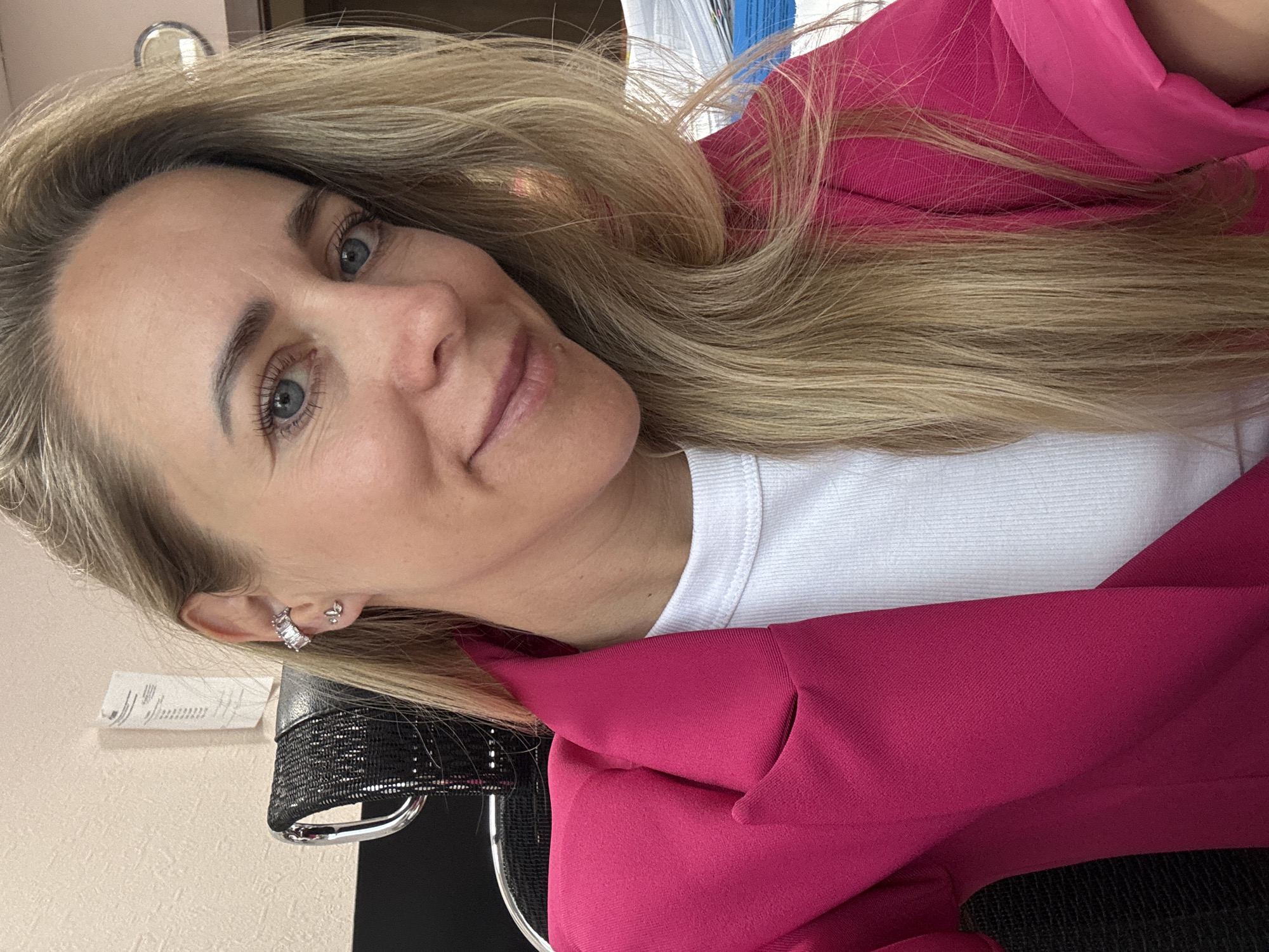 A medium shot from the chest up shows a blonde woman with blue eyes, wearing a bright pink blazer over a white t-shirt. She is looking towards the camera with a slight smile. Her hair is long and blonde, falling over her right shoulder. She is wearing a diamond-studded ear cuff on her left ear, and a small butterfly stud earring. In the background, to her left, is the dark mesh of an office chair. To her right, there is a stack of papers and a wall with a bulletin or notice board. The scene appears to be an office environment, possibly in the city of Chaikovsky, Russia.